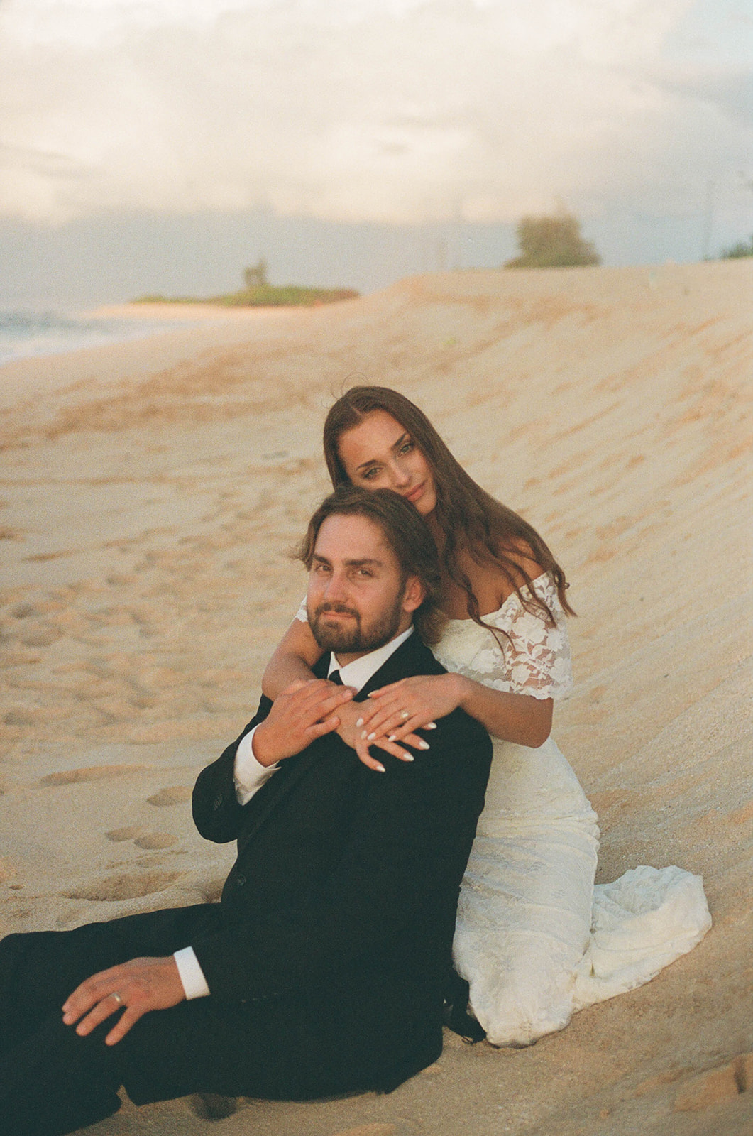 The bride sitting behind the groom as they sit on the beach hugging onto each other.