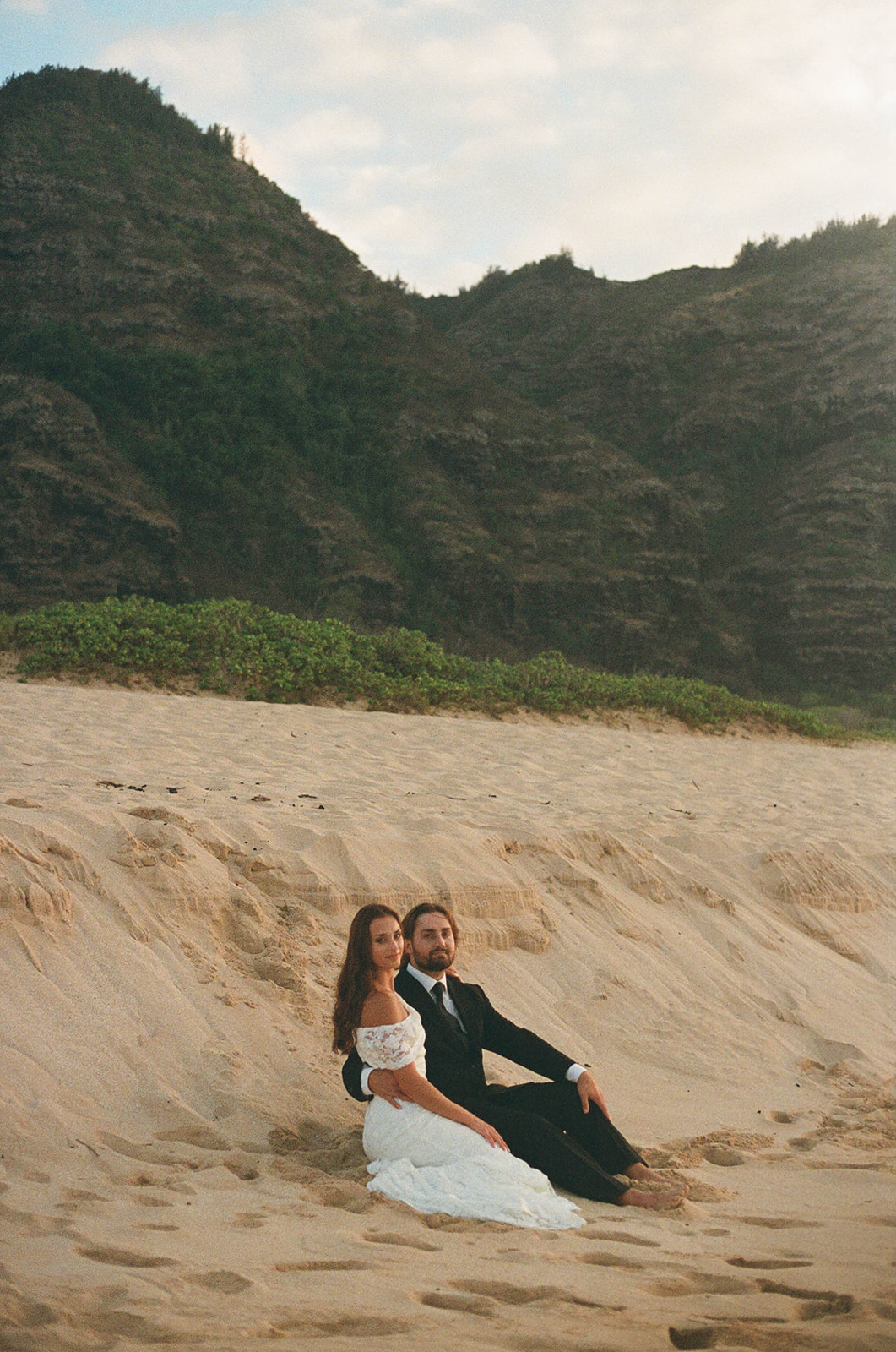 Couple sitting close together on a sandy dune overlooking the ocean, captured during their intimate Kauai Wedding Planning session photos.
