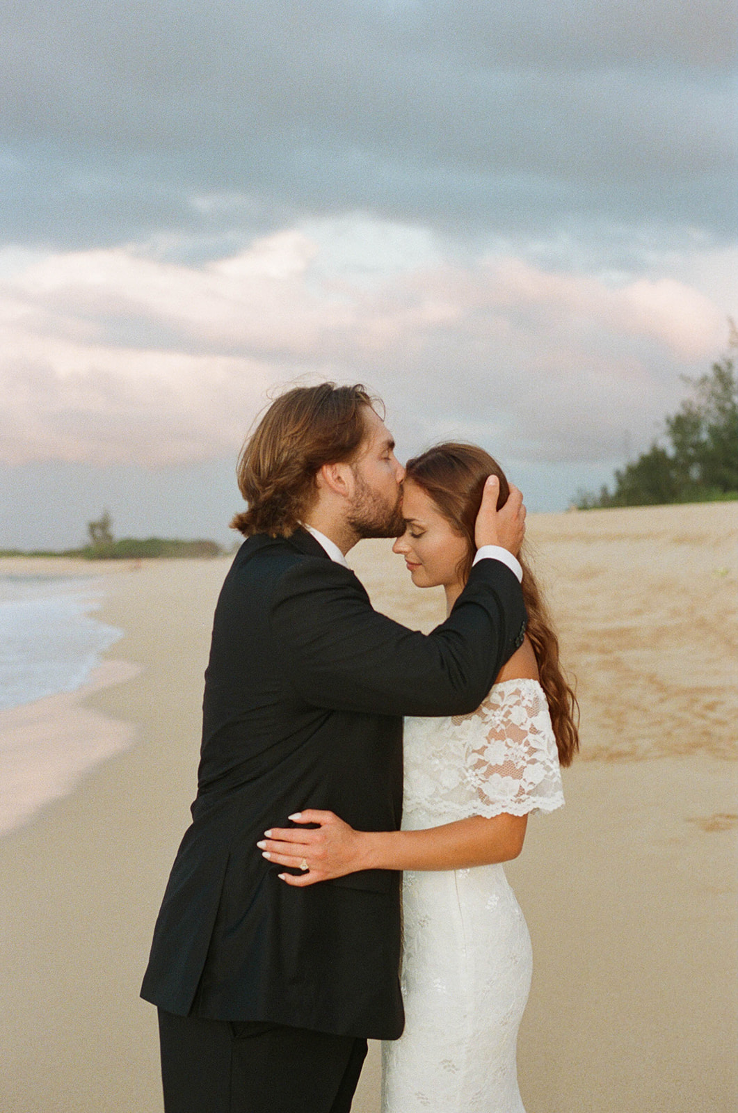 Groom kissing the bride’s forehead as they hold hands in the golden sunlight of their Kauai Wedding, surrounded by mountain views.