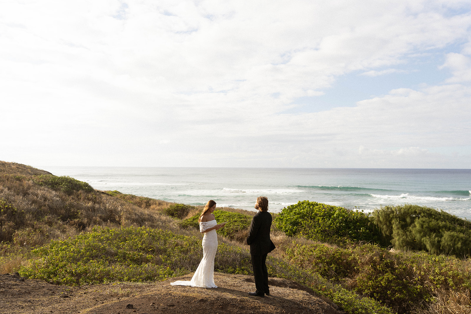 Bride and groom standing alone on a coastal overlook as they exchange vows with waves rolling in behind them, documented in a wide, atmospheric documentary wedding photography shot.