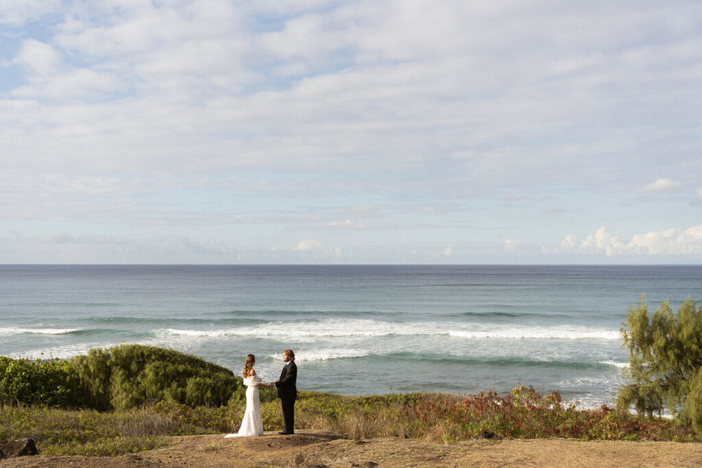 Bride and groom standing on a coastal overlook as they exchange vows with waves rolling in behind them during their Kauai wedding.