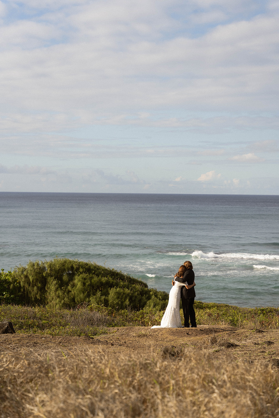 Bride and groom embracing on a grassy cliff as waves crash below them, a quiet moment from their Kauai Wedding day.