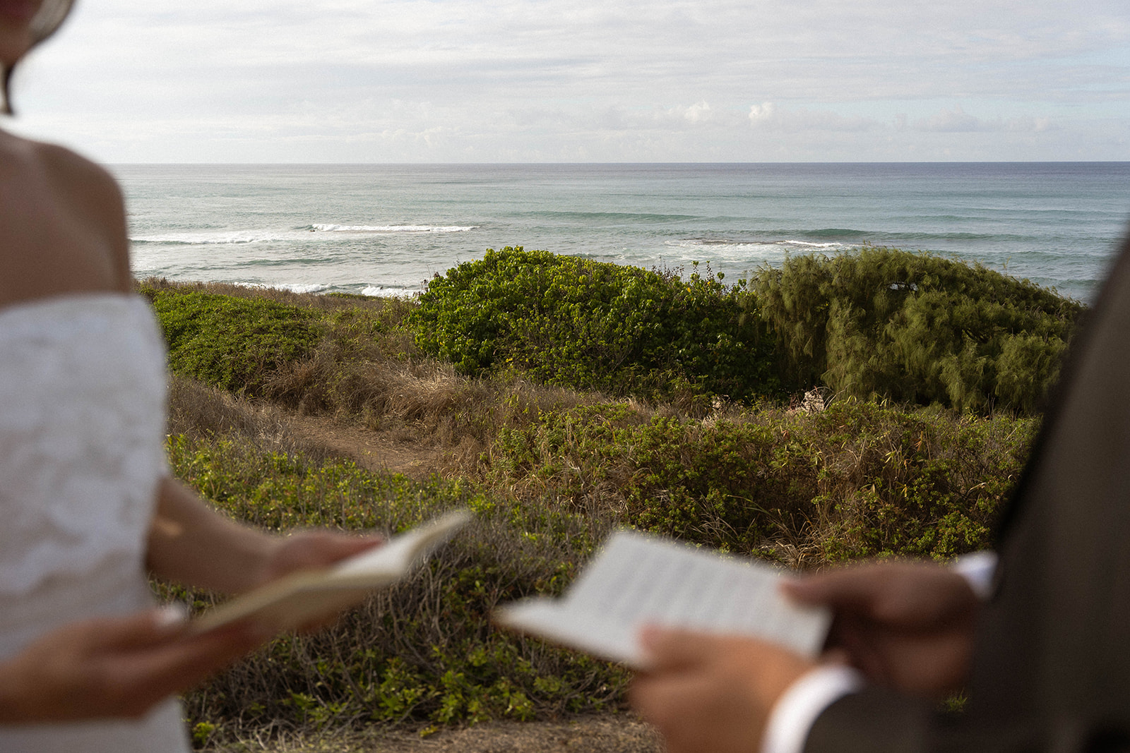 A blurred foreground of the couple reading their vows with the ocean behind them, highlighting the scenery and intimacy in a quiet documentary wedding photography perspective.