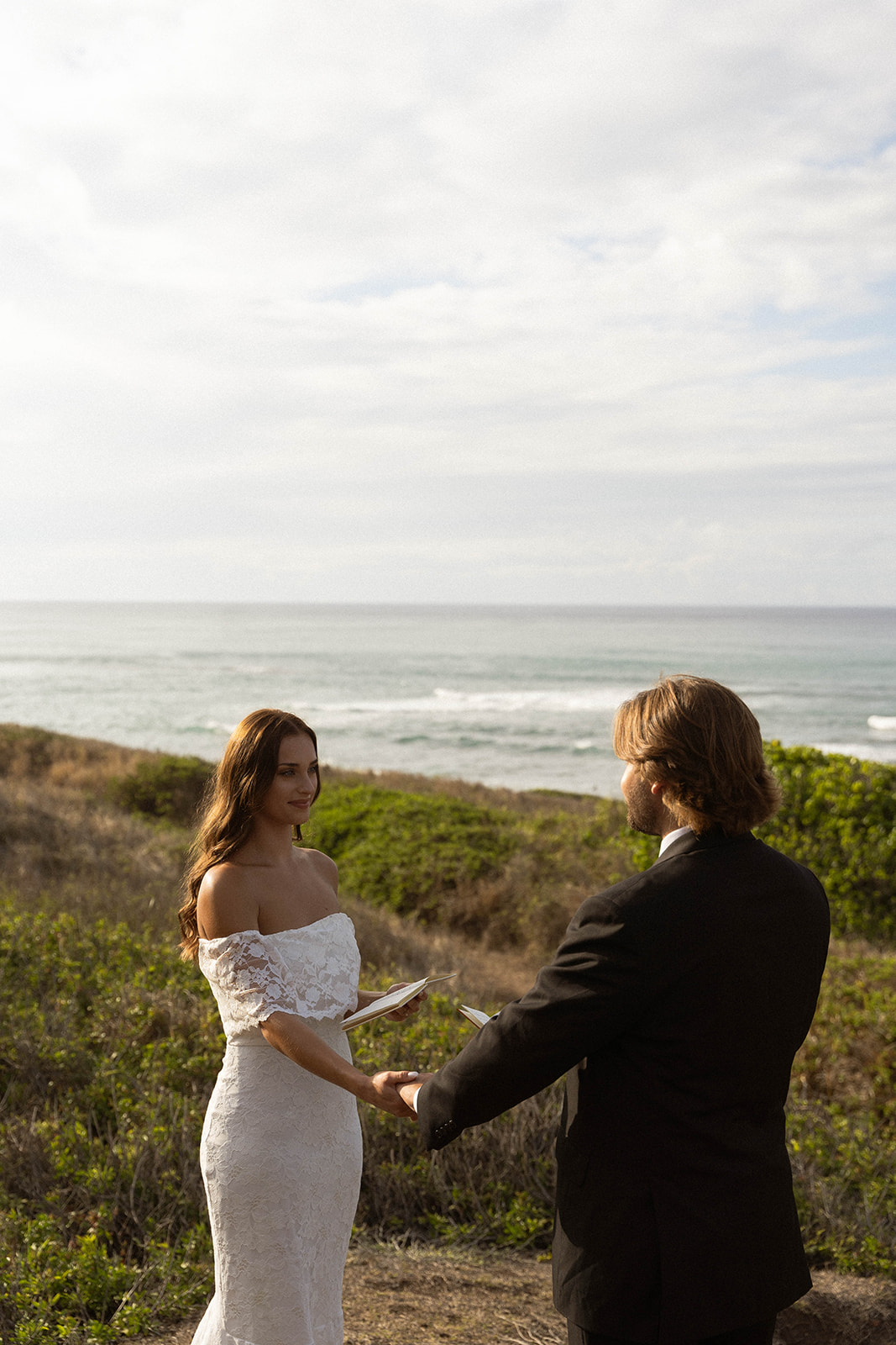 Bride holding a vow book and smiling softly at her groom during an intimate cliffside ceremony, photographed with a natural documentary wedding photography approach.