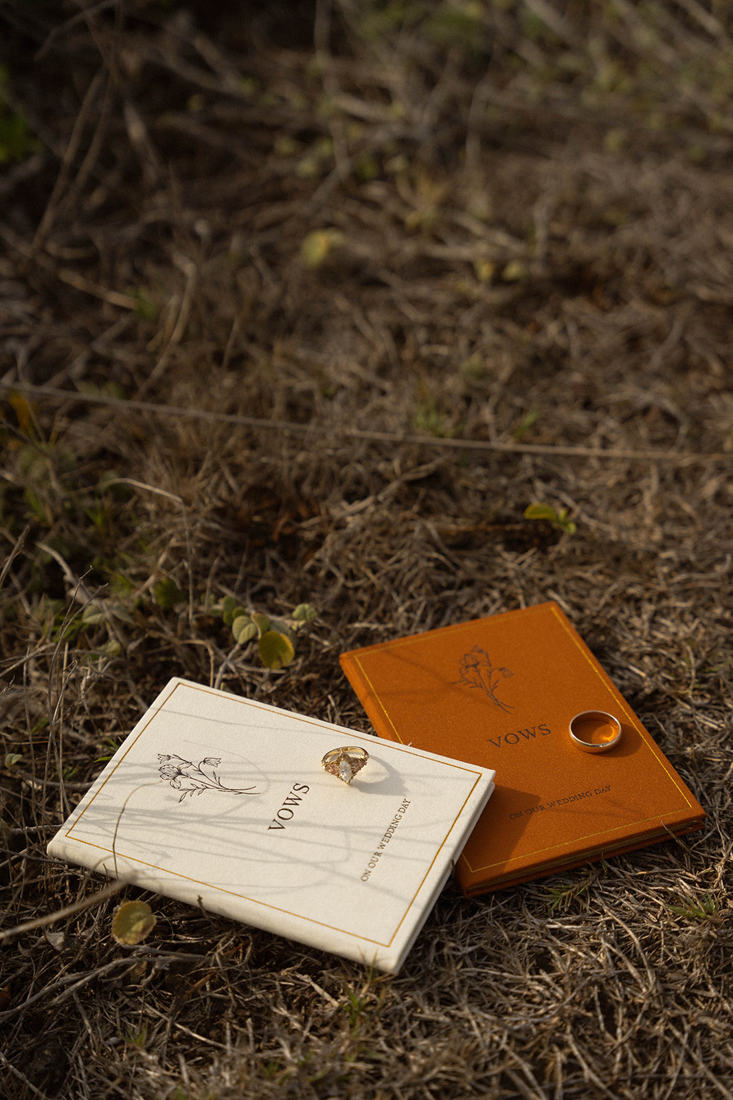 Bride and groom’s vow books resting on dry grass with their rings placed on top, captured during their elegant Kauai Wedding details.