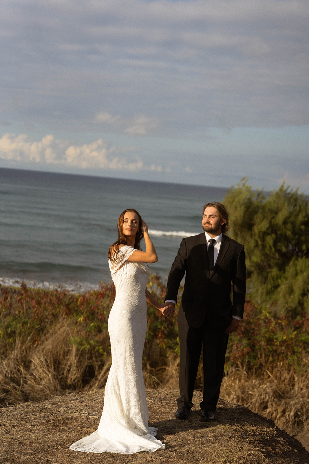 Bride brushing her hair back while holding hands with the groom on a coastal cliff during their Kauai Wedding Planning session.