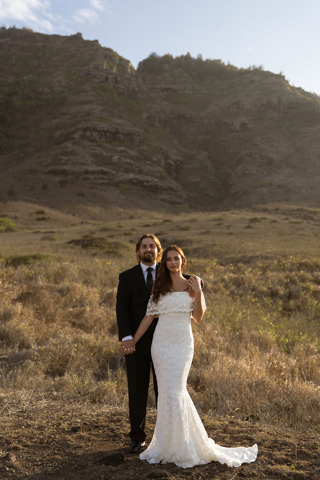 The couple stands together in an open field with mountains behind them, holding hands and standing close in warm sunlight, captured in a peaceful documentary wedding photography style.