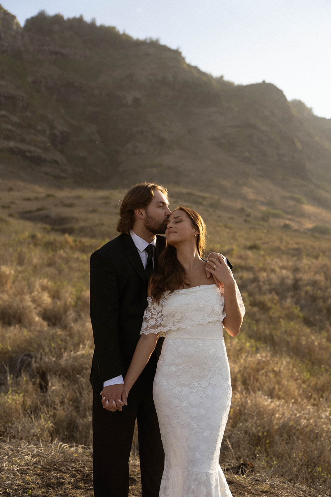 Groom kissing the bride’s forehead as they hold hands in the golden sunlight of their Kauai Wedding, surrounded by mountain views.