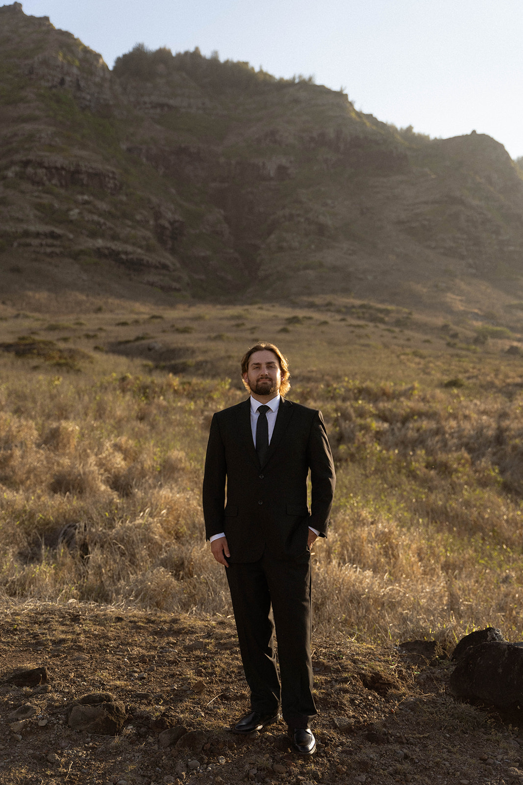 The groom stands alone against a mountain backdrop in soft afternoon light, wearing a black suit and tie, photographed in a clean and minimal documentary wedding photography style.