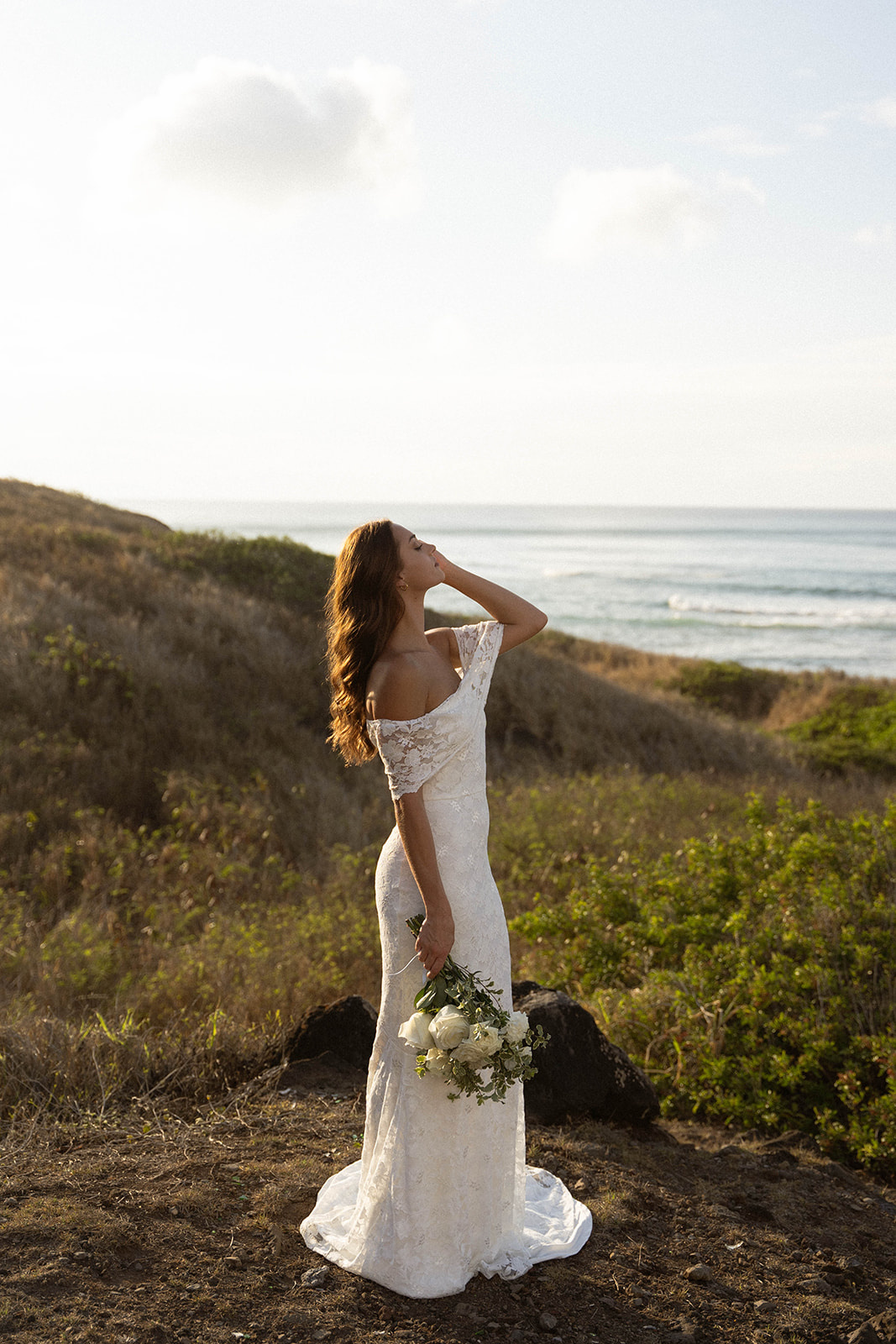 The bride stands facing the ocean with golden light hitting her hair, holding her bouquet at her side as she looks upward, documented in a serene documentary wedding photography moment.