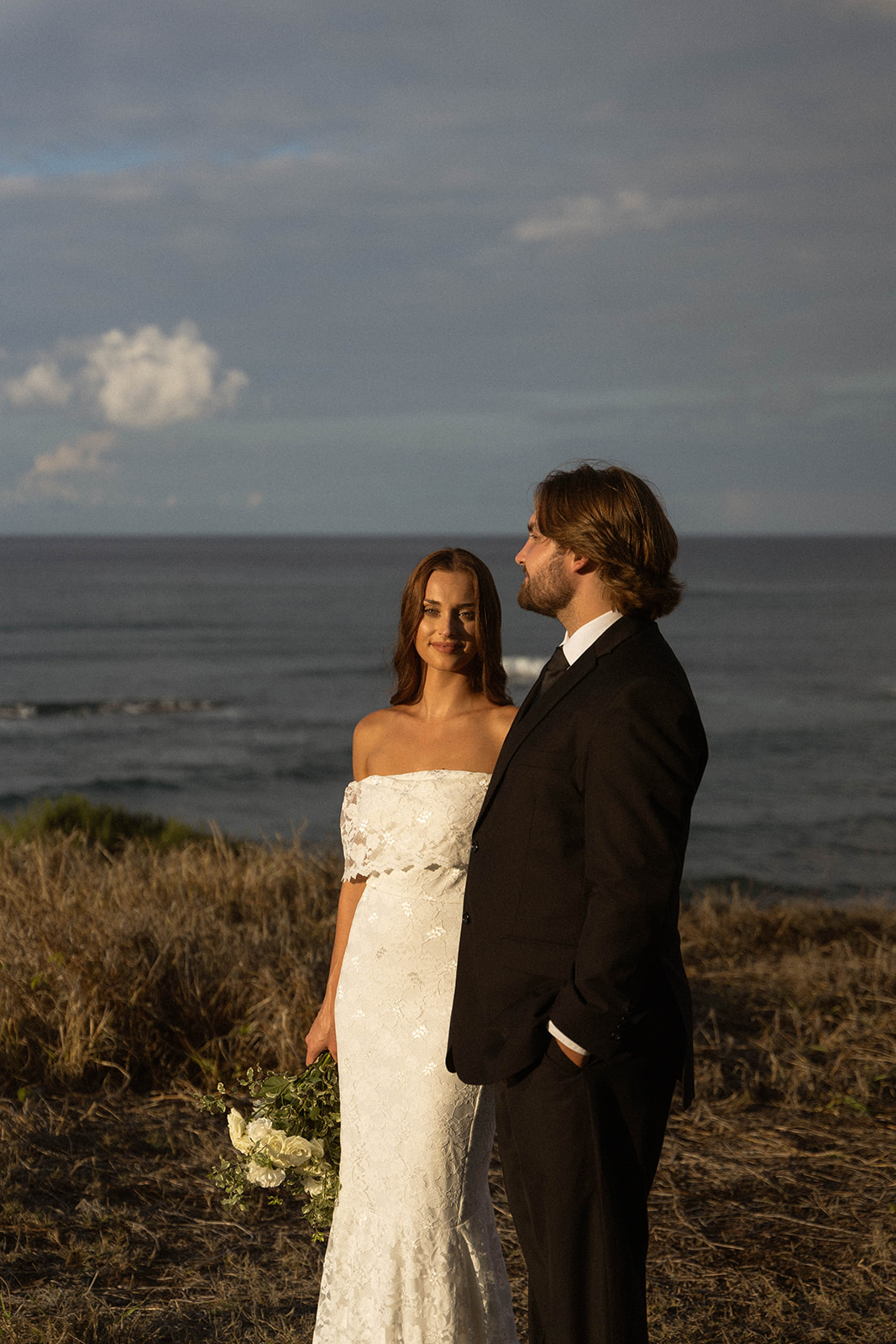 The bride and groom stand together by the coastline with warm golden light on their faces, the bride holding her bouquet while the groom looks out toward the ocean, photographed in a calm documentary wedding photography style.