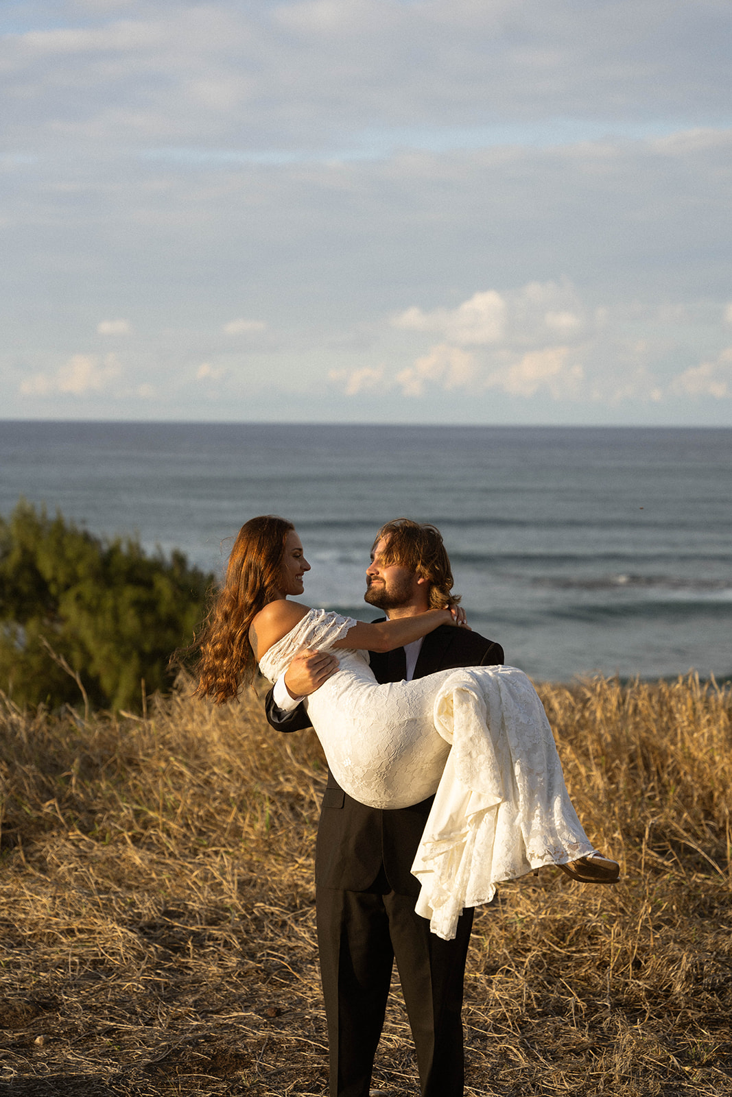The groom lifts the bride in his arms on a cliffside overlooking the ocean, both smiling toward each other as the sun sets, captured in joyful documentary wedding photography fashion.
