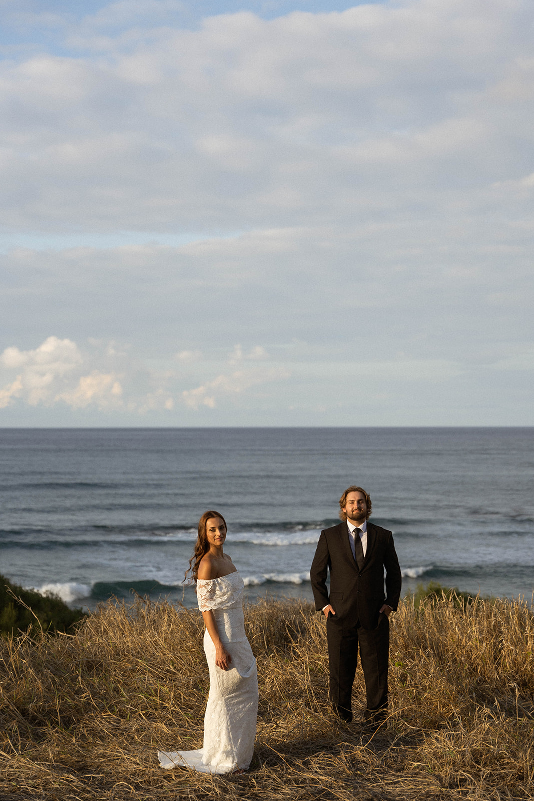 Bride and groom standing apart on a grassy cliffside overlooking the ocean, photographed during their Kauai Wedding Planning session.