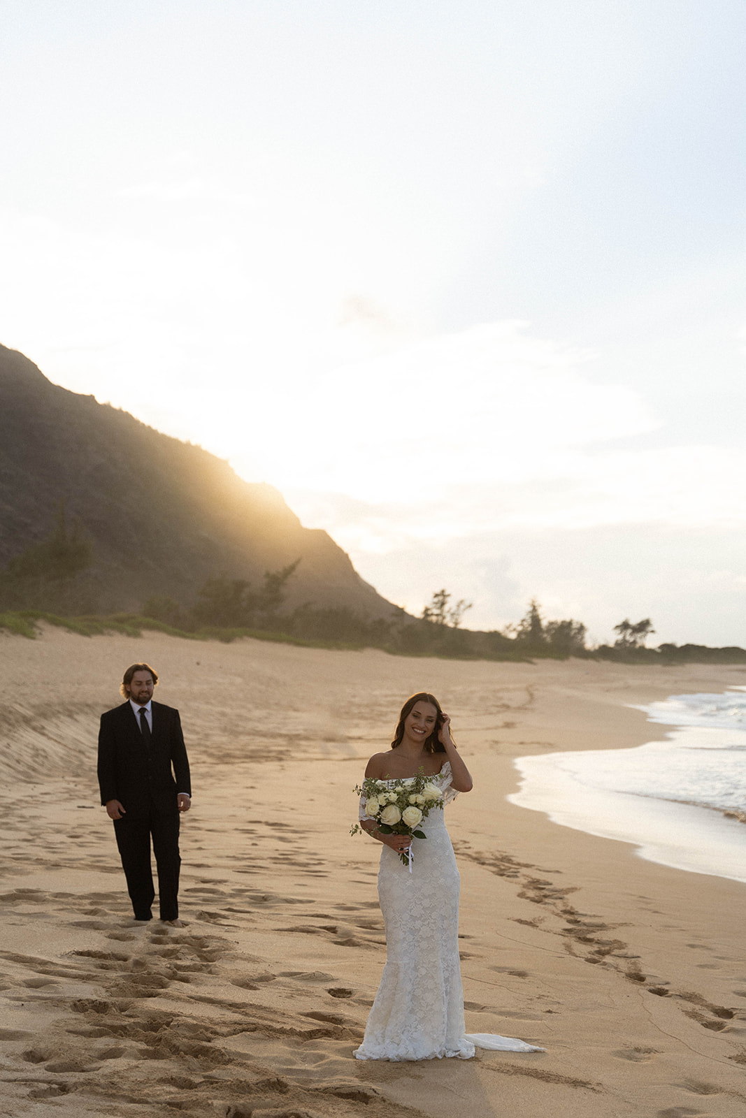 Bride walking along the beach with her bouquet while the groom follows behind her, illuminated by soft sunrise light in true documentary wedding photography style.