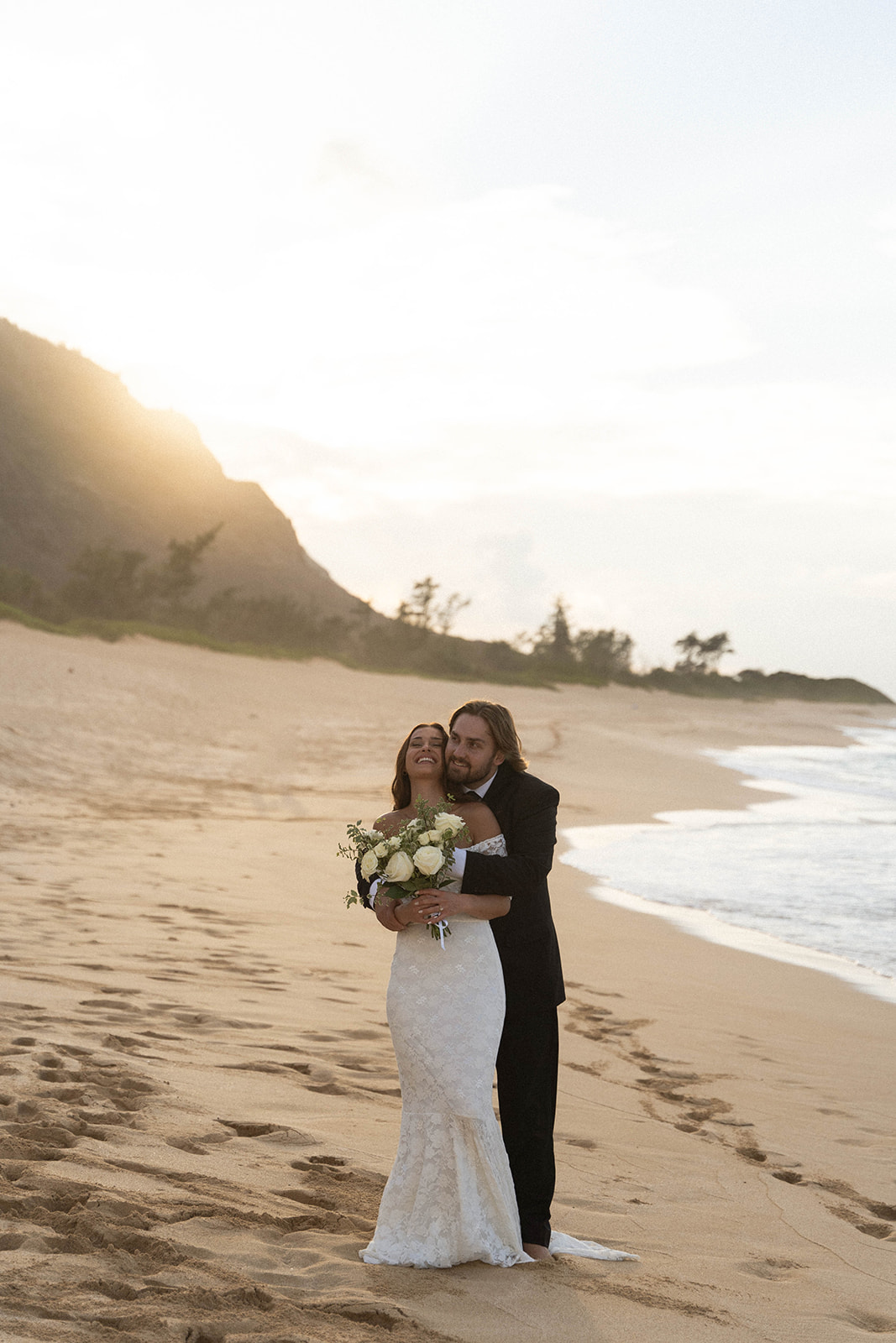 A couple embraces on a quiet beach at sunset, the bride holding white roses and laughing as her partner wraps his arms around her from behind, captured in a soft and natural documentary wedding photography style.