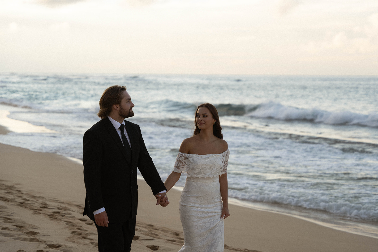 Bride and groom holding hands while walking barefoot along a quiet shoreline during their Kauai Wedding, with waves rolling in behind them at sunset.