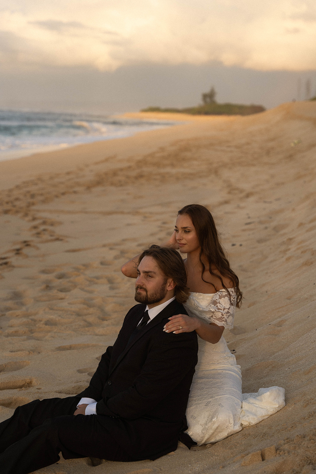 Bride sitting behind the groom on the sand with her hand gently resting on his shoulder, a quiet and relaxed moment framed in a natural documentary wedding photography style.