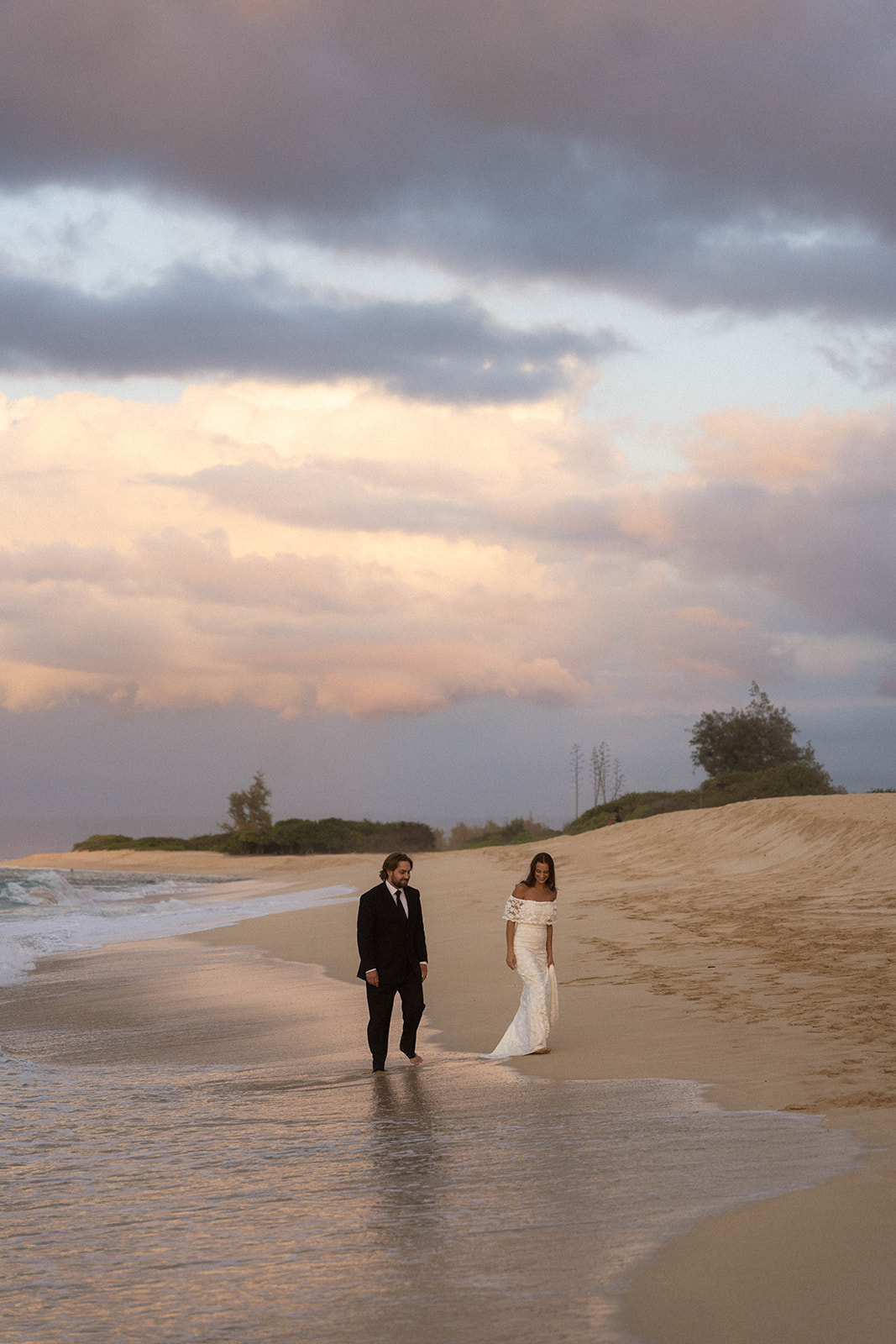 Bride and groom walking beside the water as pastel clouds fill the sky, a peaceful scene from their Kauai Wedding Planning journey.