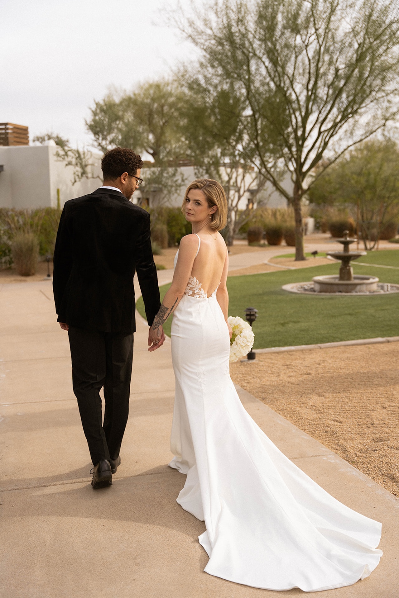 Couple standing hand in hand on a modern walkway, the bride’s fitted gown flowing behind her.