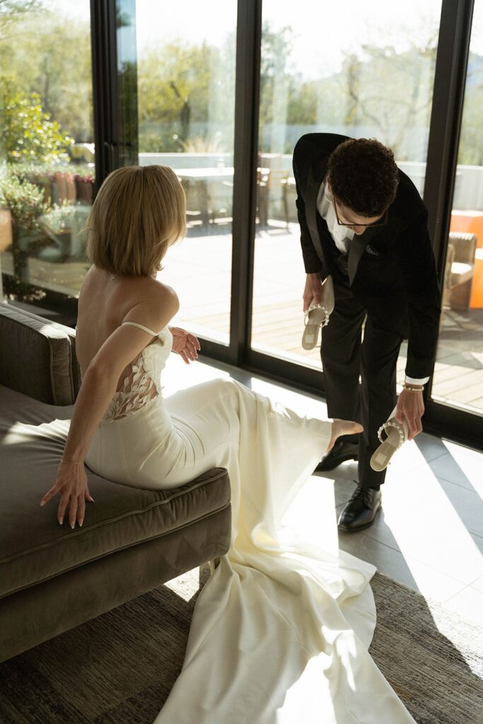 Groom helping bride with her heels beside the glass patio doors, soft afternoon light filling the space.