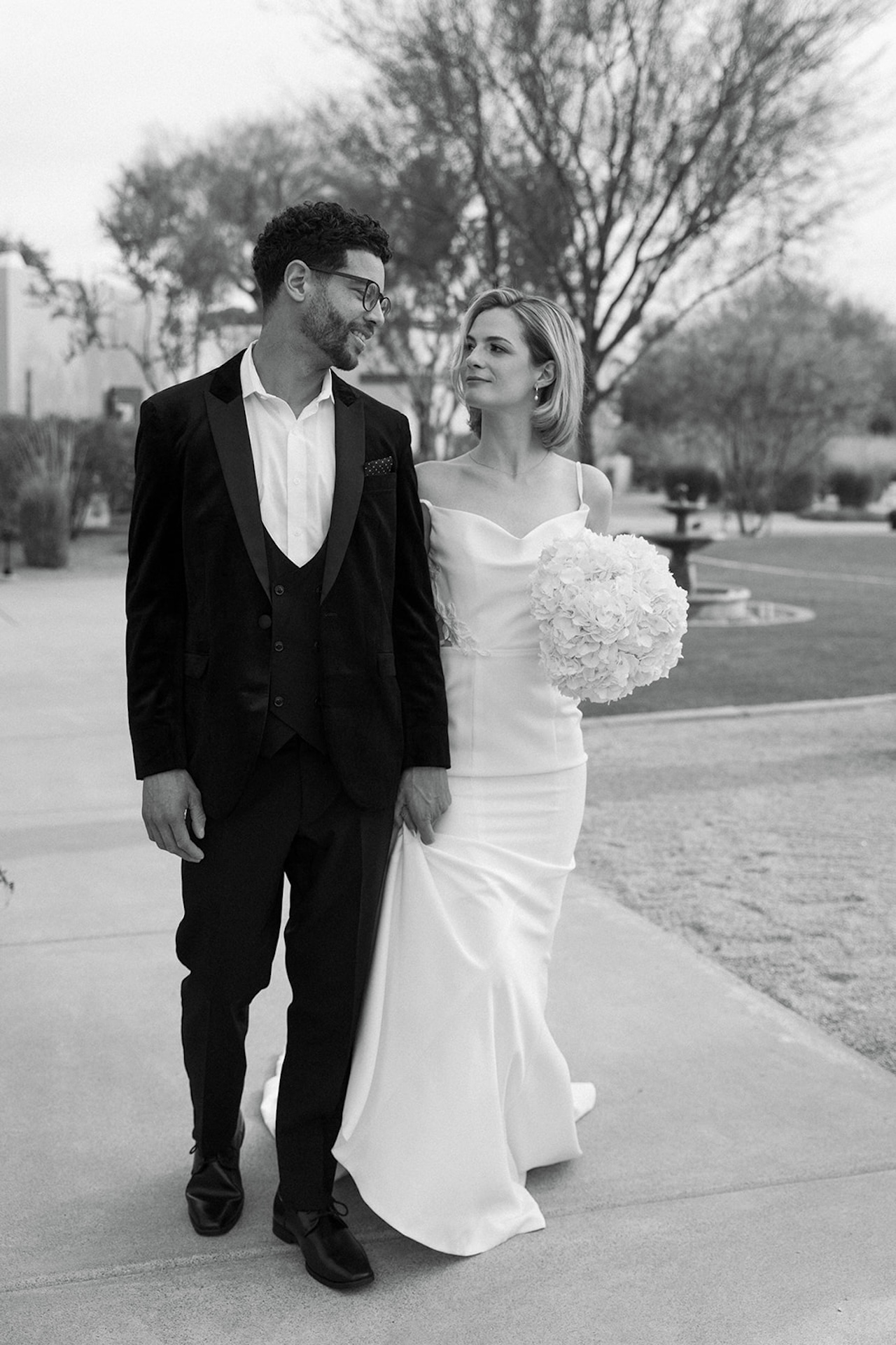 Black and white photo of the couple walking hand in hand, smiling and looking at each other.