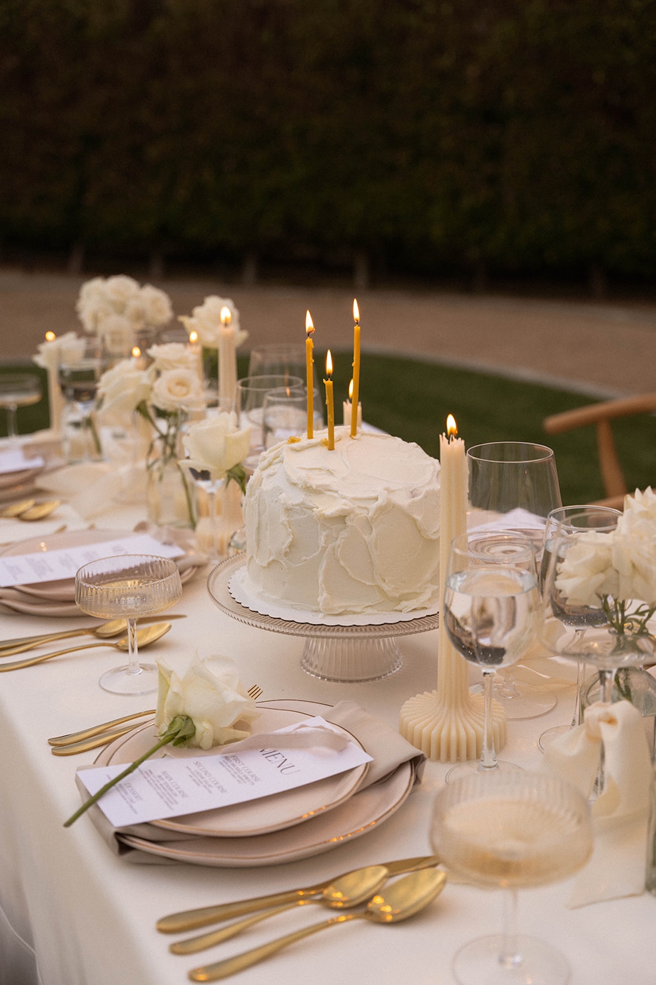 White cake with golden candles glowing among crystal glassware and roses on the dinner table.