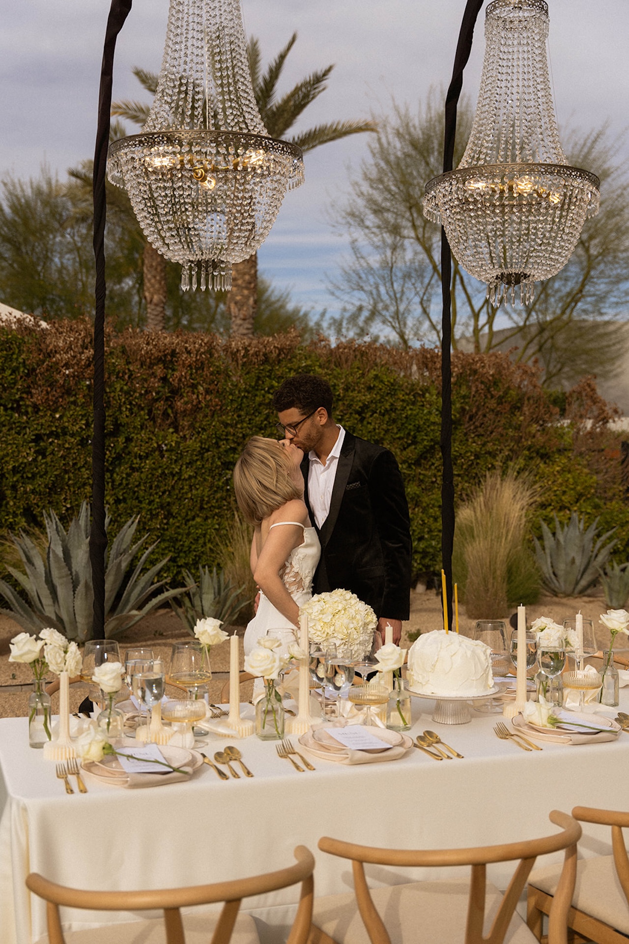 Couple sharing a quiet kiss beside the reception table set with candles and florals under crystal chandeliers.