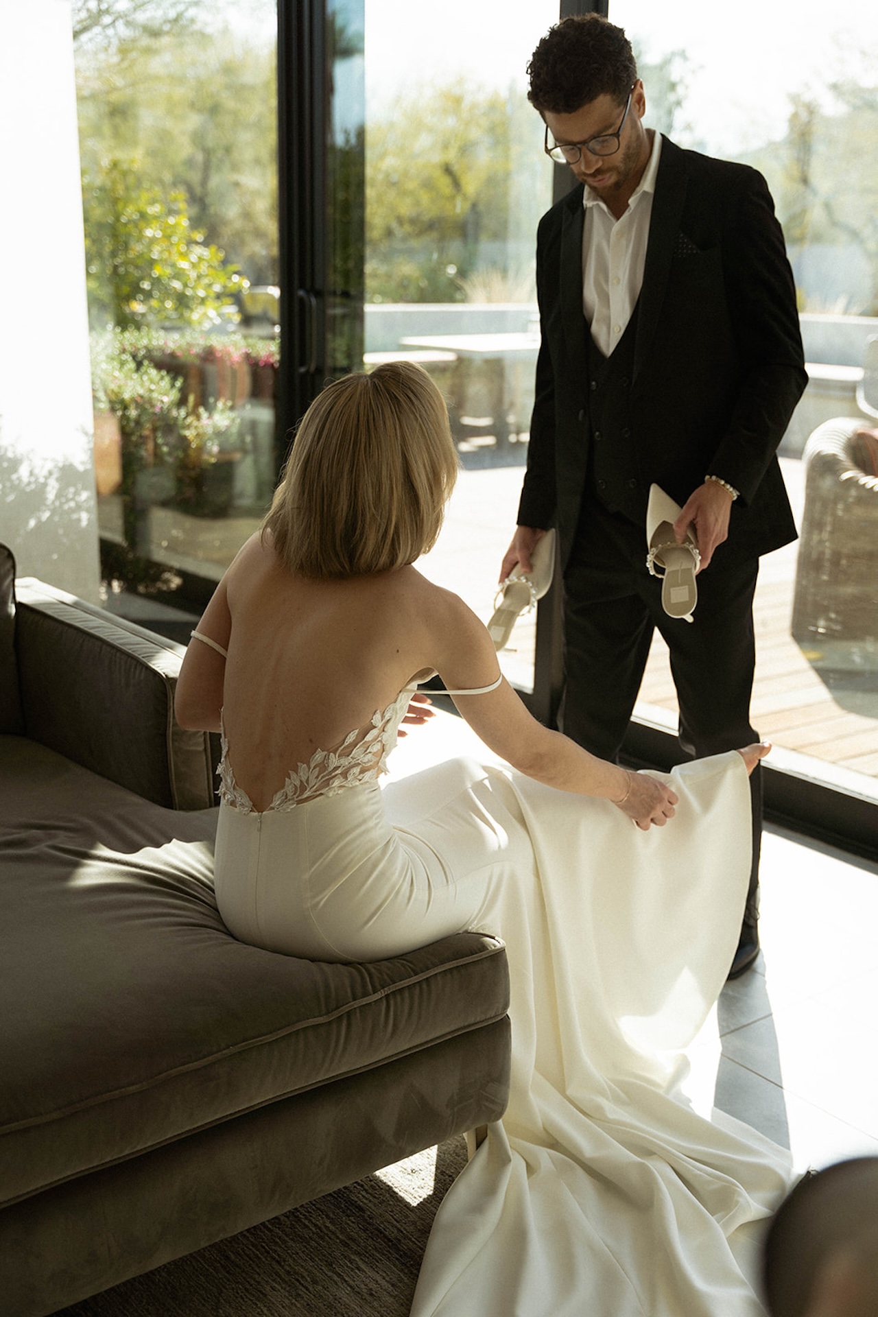 Groom holding bride’s shoes as she sits on a sofa, light streaming through modern glass doors.