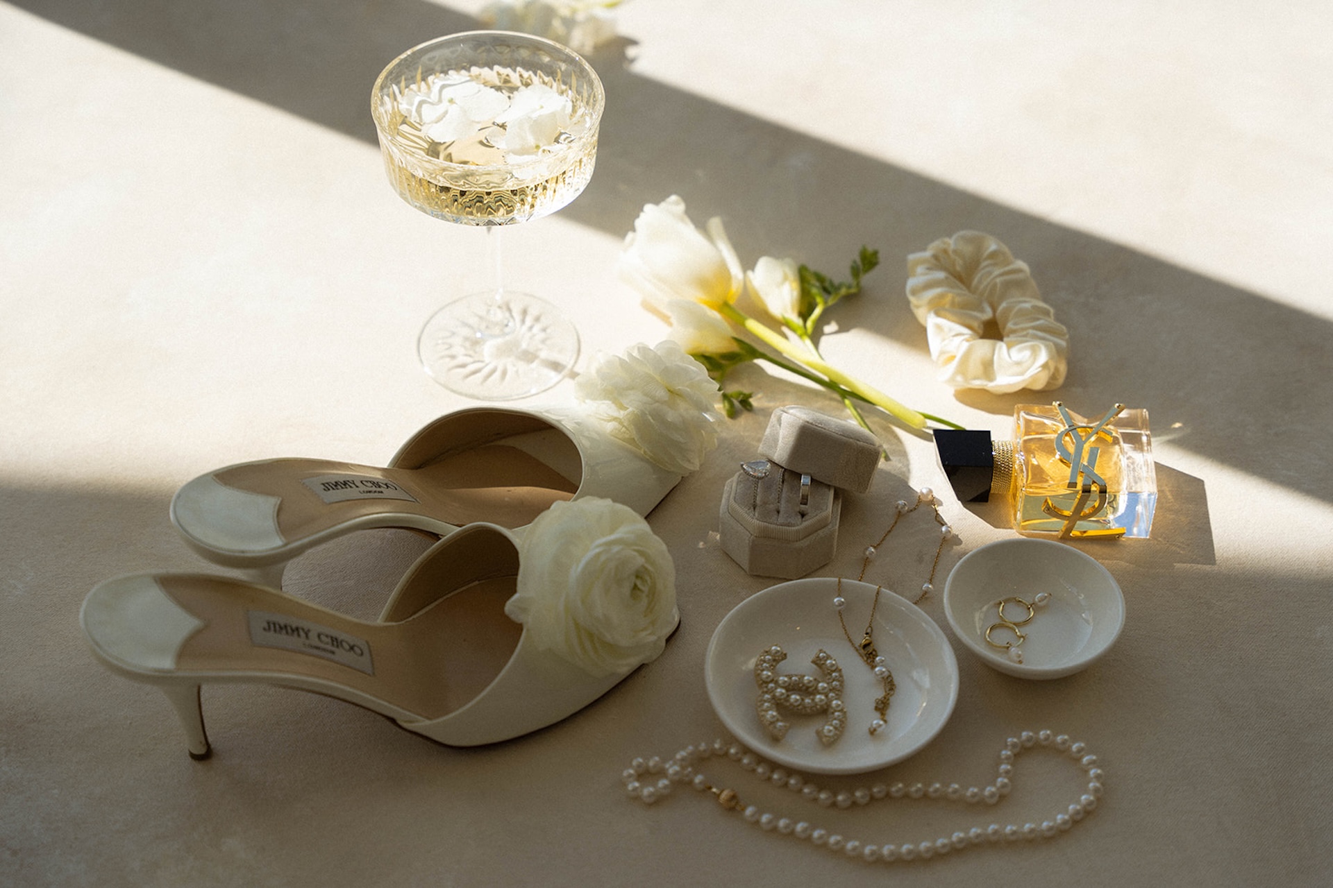 Flat lay of bridal details including Jimmy Choo heels, pearl jewelry, perfume, and champagne glass.