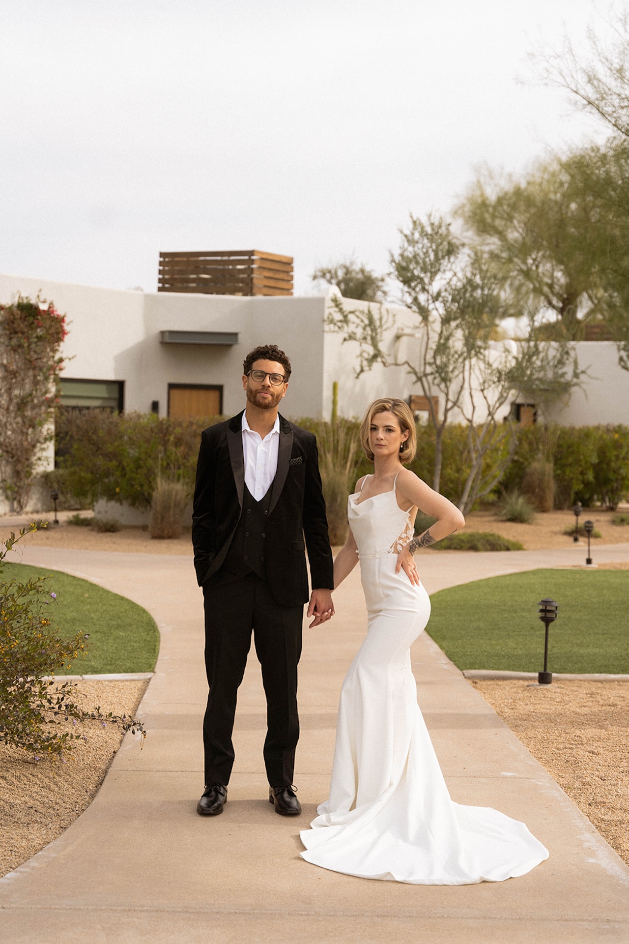 Couple posing confidently outdoors, surrounded by desert greenery and modern architecture.