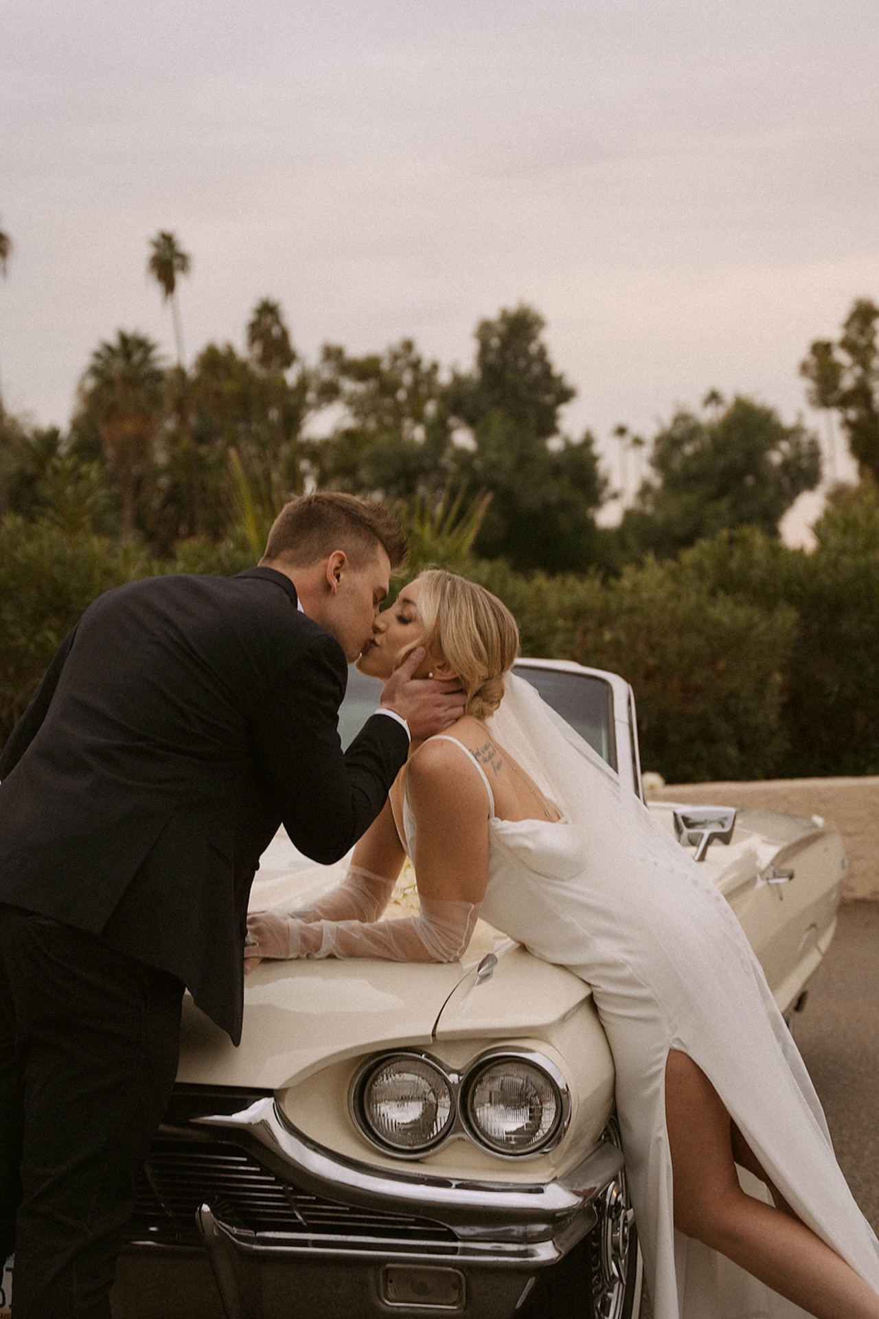 Bride and groom leaning over the hood of a convertable and sharing a kiss with palm trees in the background during their intimate wedding photos.