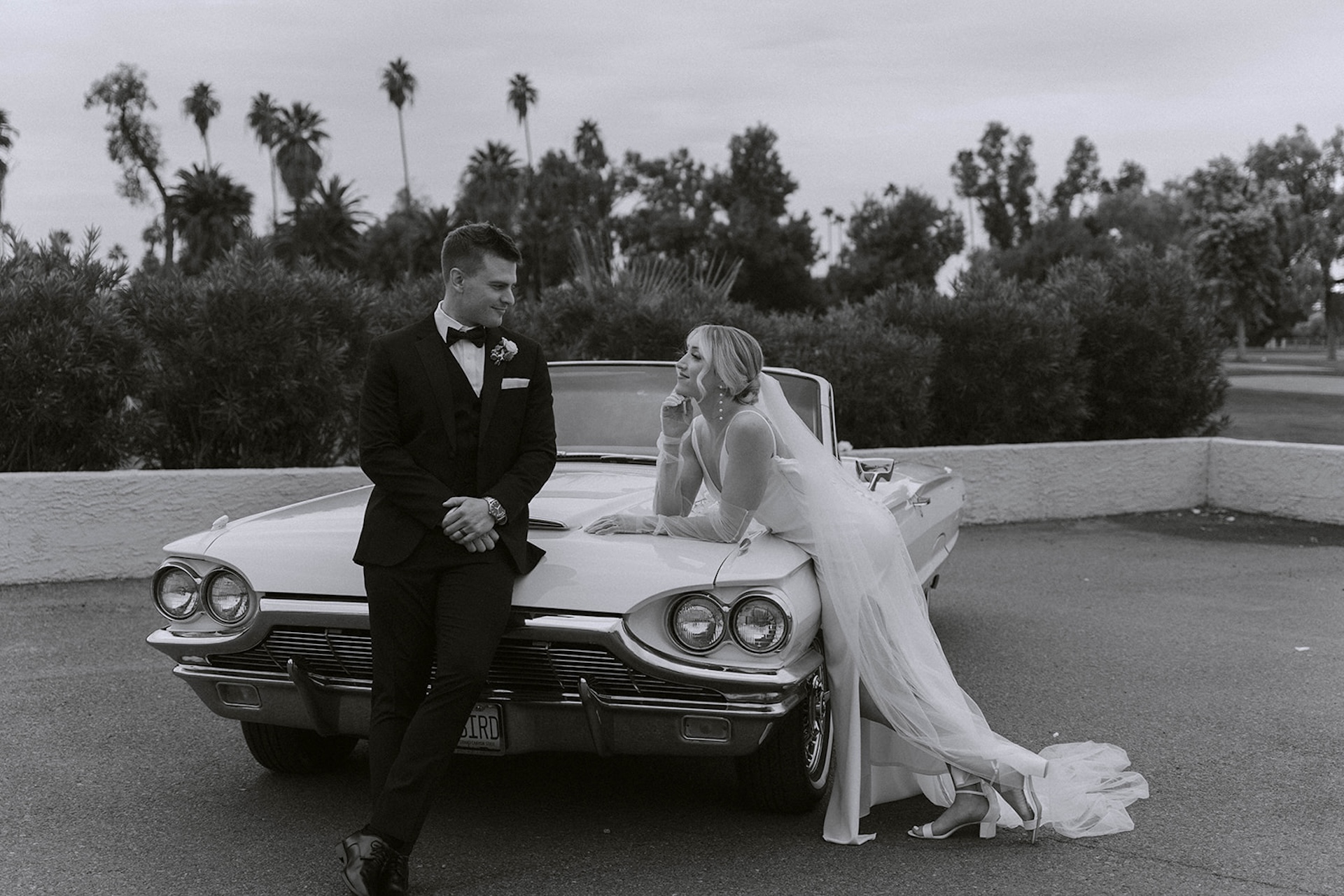 Bride and groom leaning against a vintage white convertible on their wedding day.
