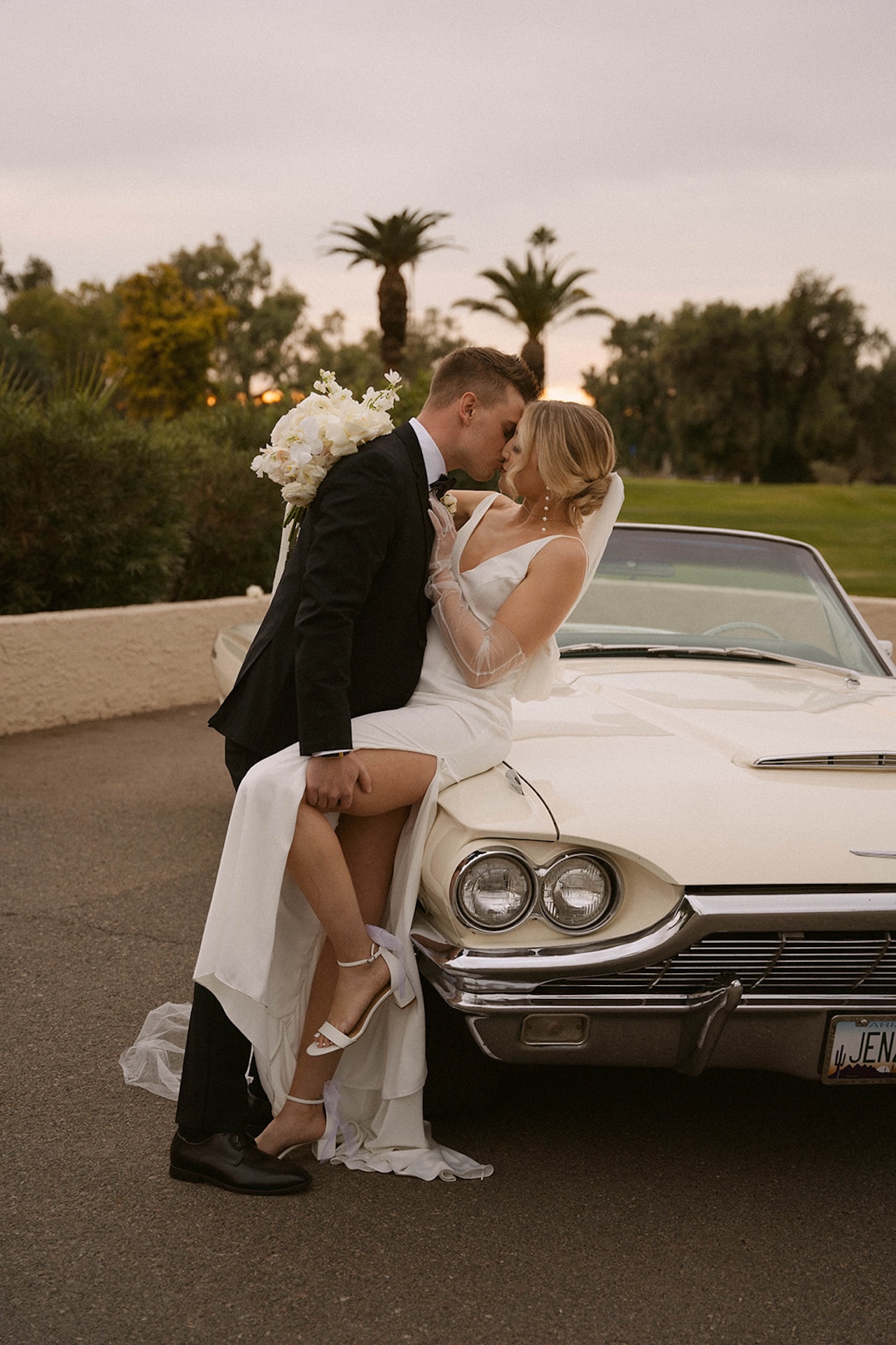 Stunning bride and groom portrait in Malibu. The bride and groom are leaning against a yellow classic convertable as he slightly dips her and holds her leg as they share a kiss with the palms behind them.