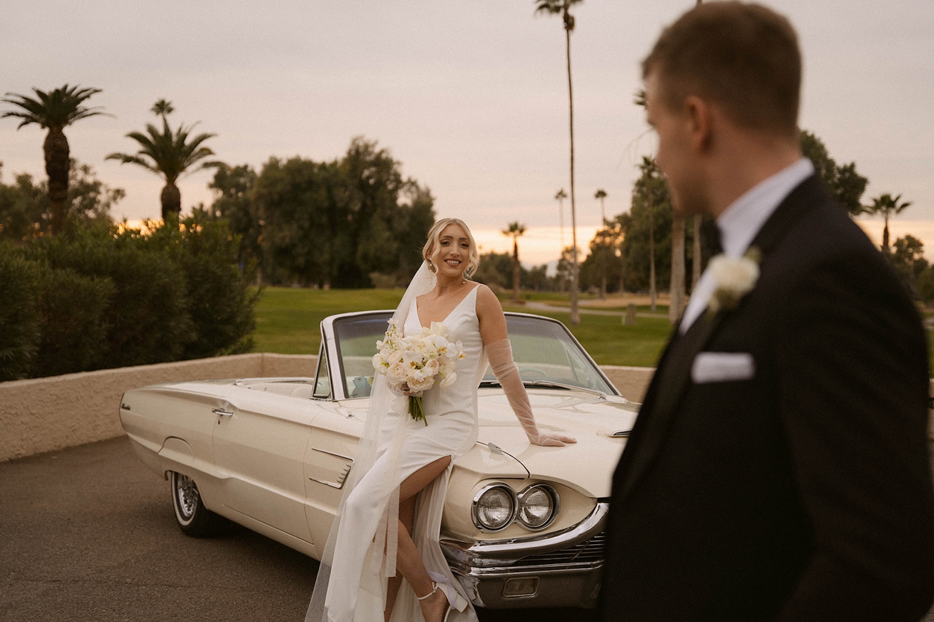 Bride leaning on a convertable looking looking at the groom as she holds her white bridal bouquet.