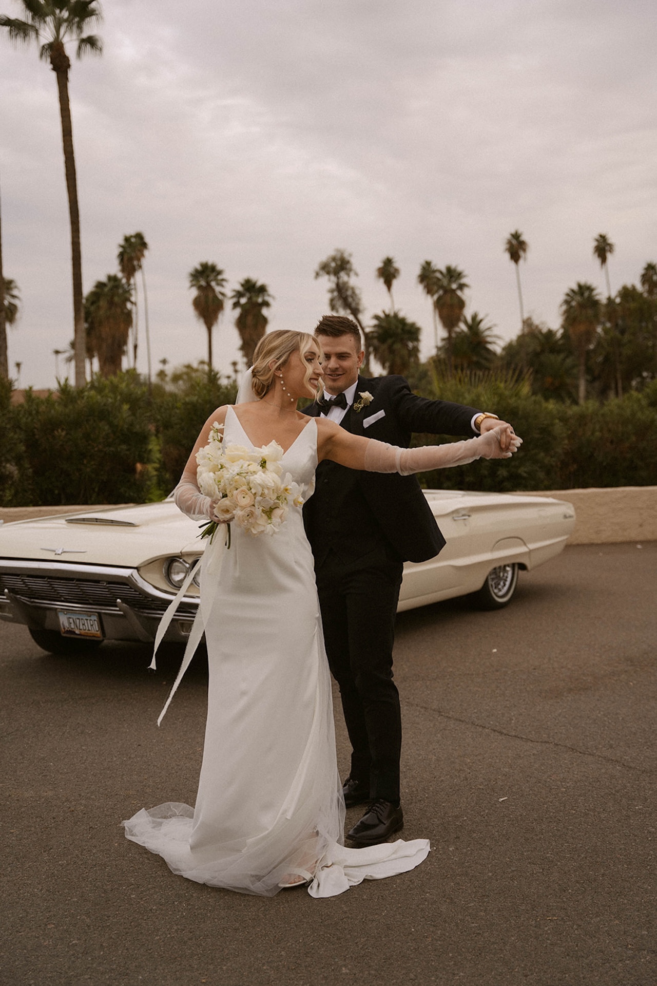 Bride and groom dancing together beside a classic car.