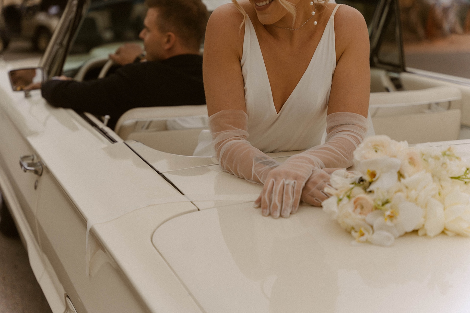 Bride smiling and leaning on the back of a white convertible with her bouquet nearby.