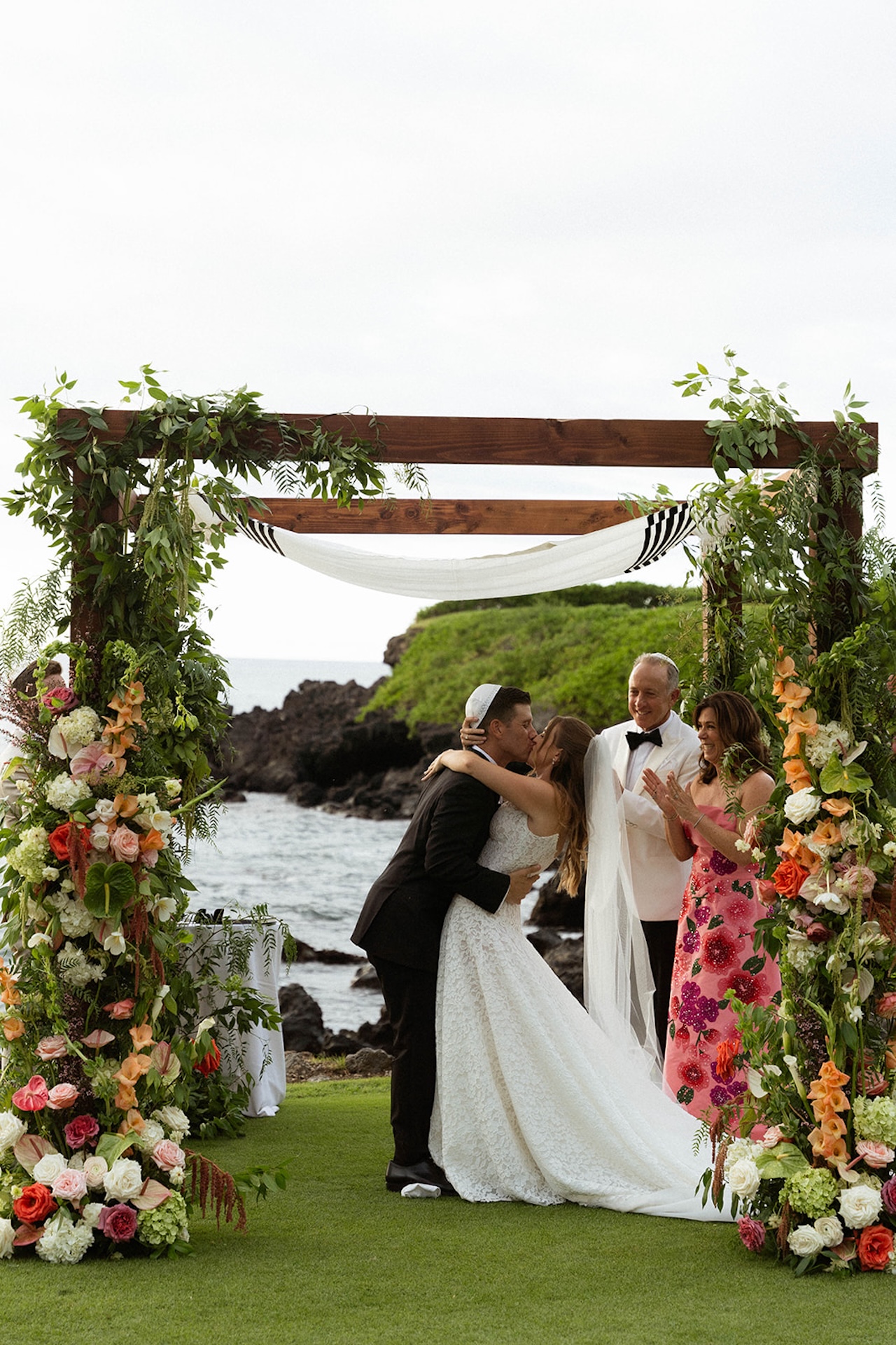 A couple shares their first kiss beneath a floral ceremony arch overlooking the ocean, a timeless moment captured during big island wedding planning.