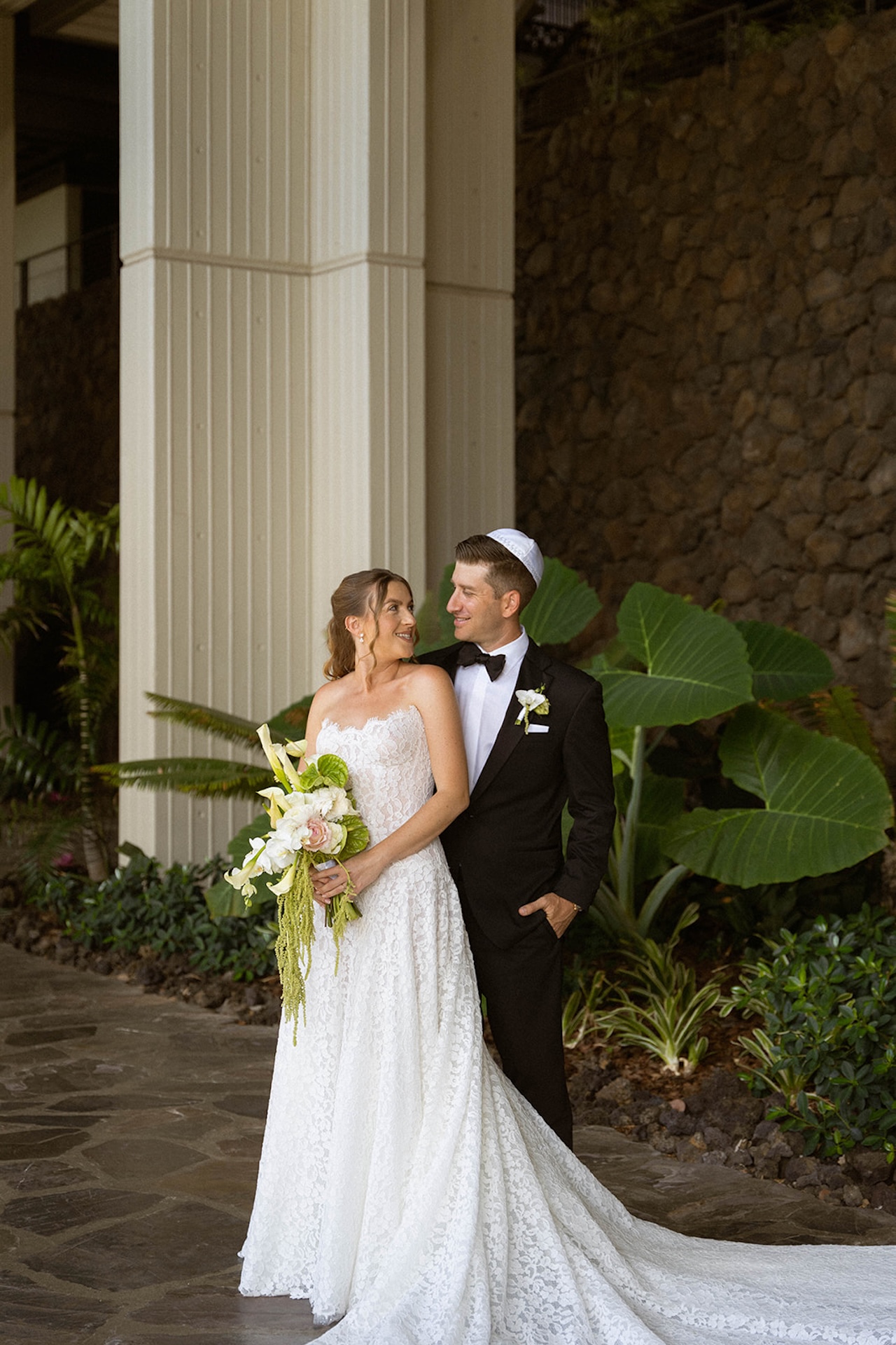 The bride and groom smile at each other while standing beneath tall columns and tropical plants at their venue.