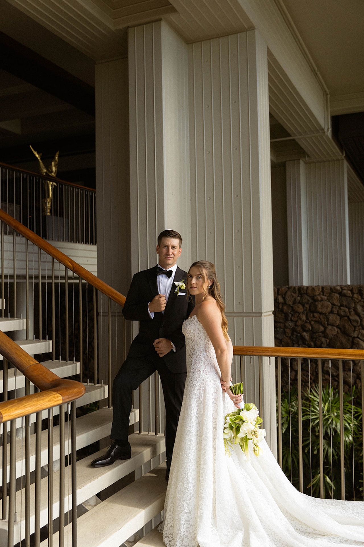The couple stands together on a modern stairway holding florals, a clean and elegant moment from Big Island wedding planning portraits.