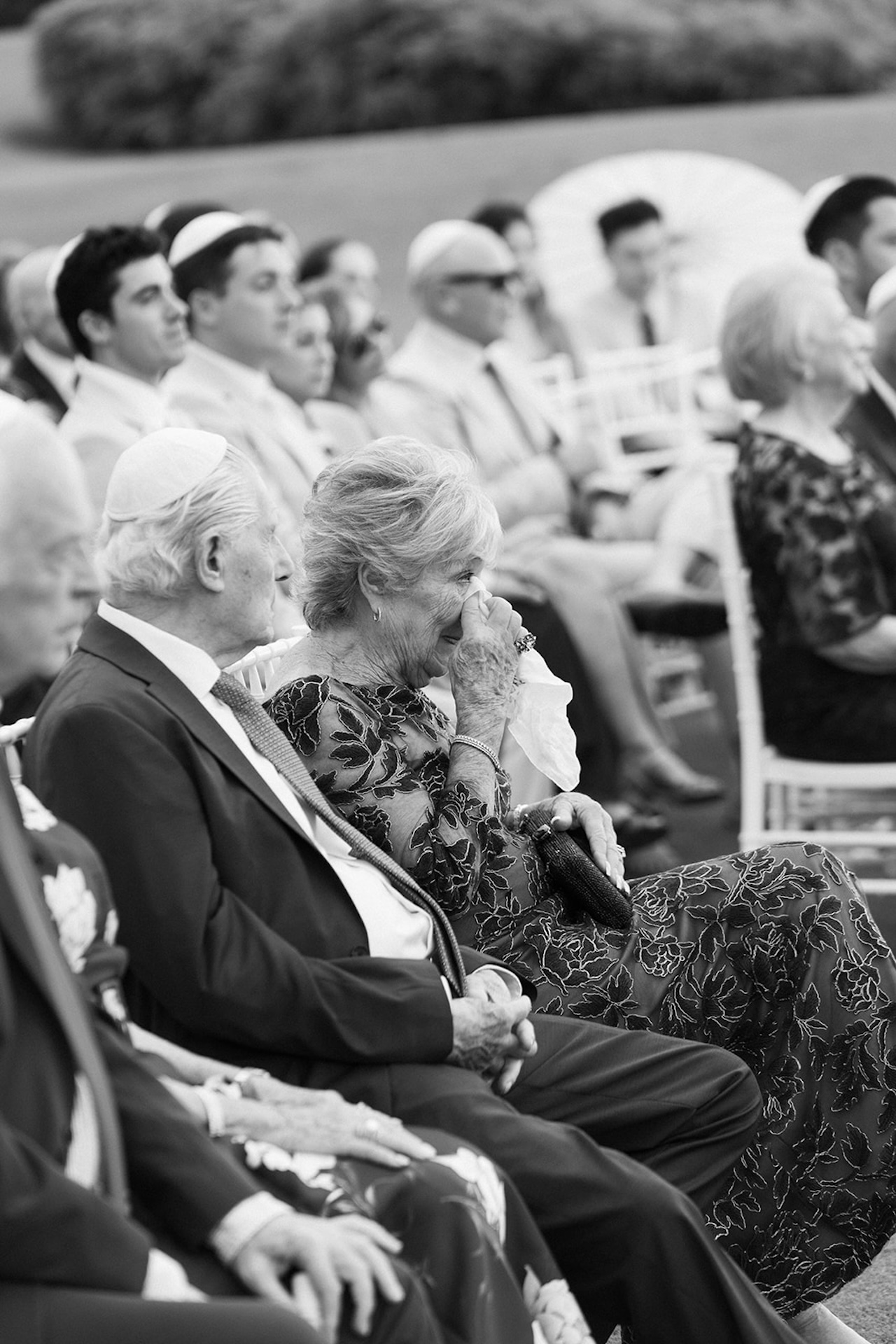 An emotional guest wipes away tears while seated during an outdoor wedding ceremony