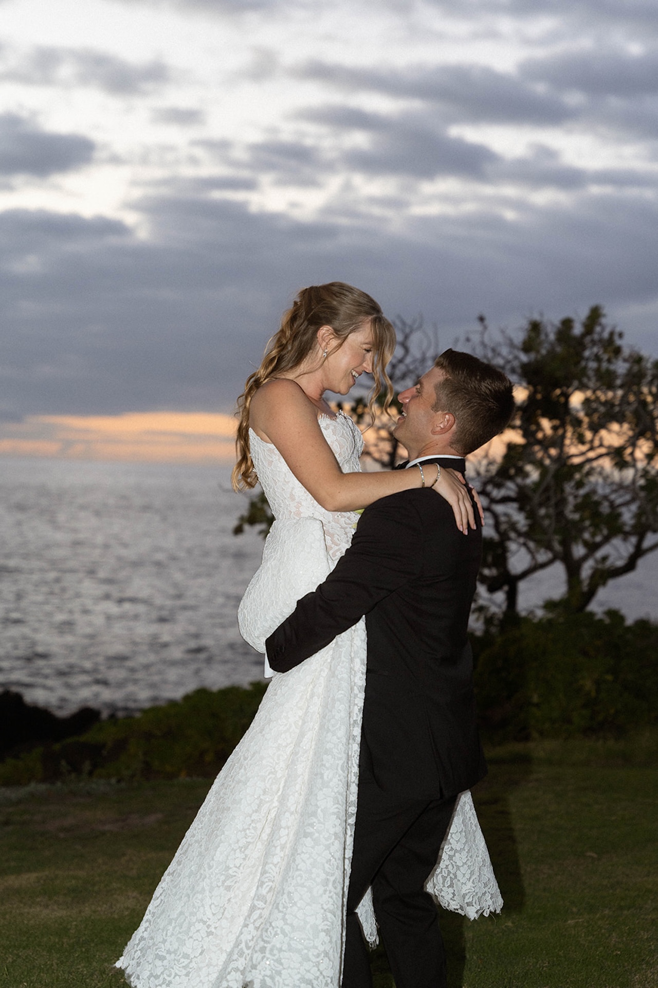 A bride and groom share a romantic lift near the ocean at sunset, a timeless portrait inspired by Big Island wedding planning.