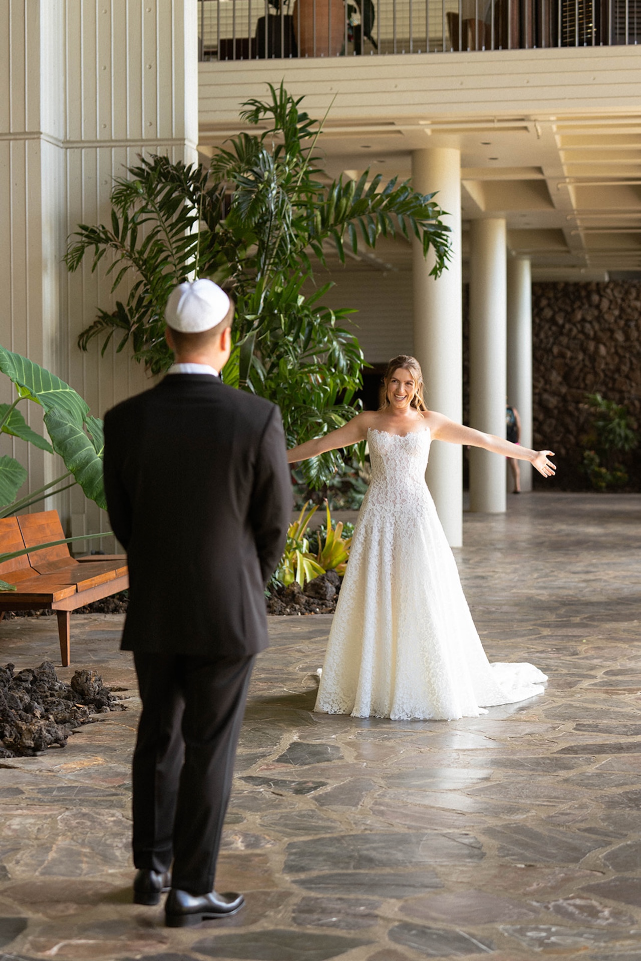 The bride opens her arms wide as she sees her partner for the first look inside a tropical resort space.