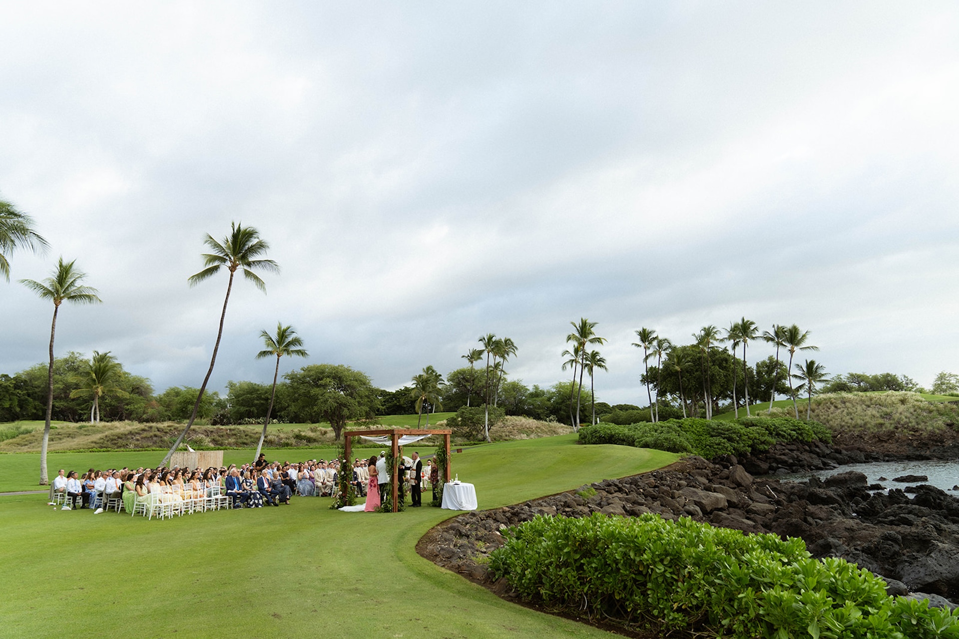 Wide view of a lush outdoor wedding ceremony setup with guests seated on green grass near the coastline.