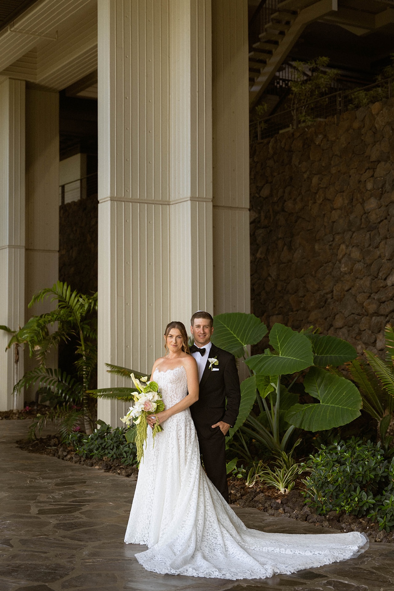 The bride and groom pose together beneath tall columns and lush tropical plants at their venue.