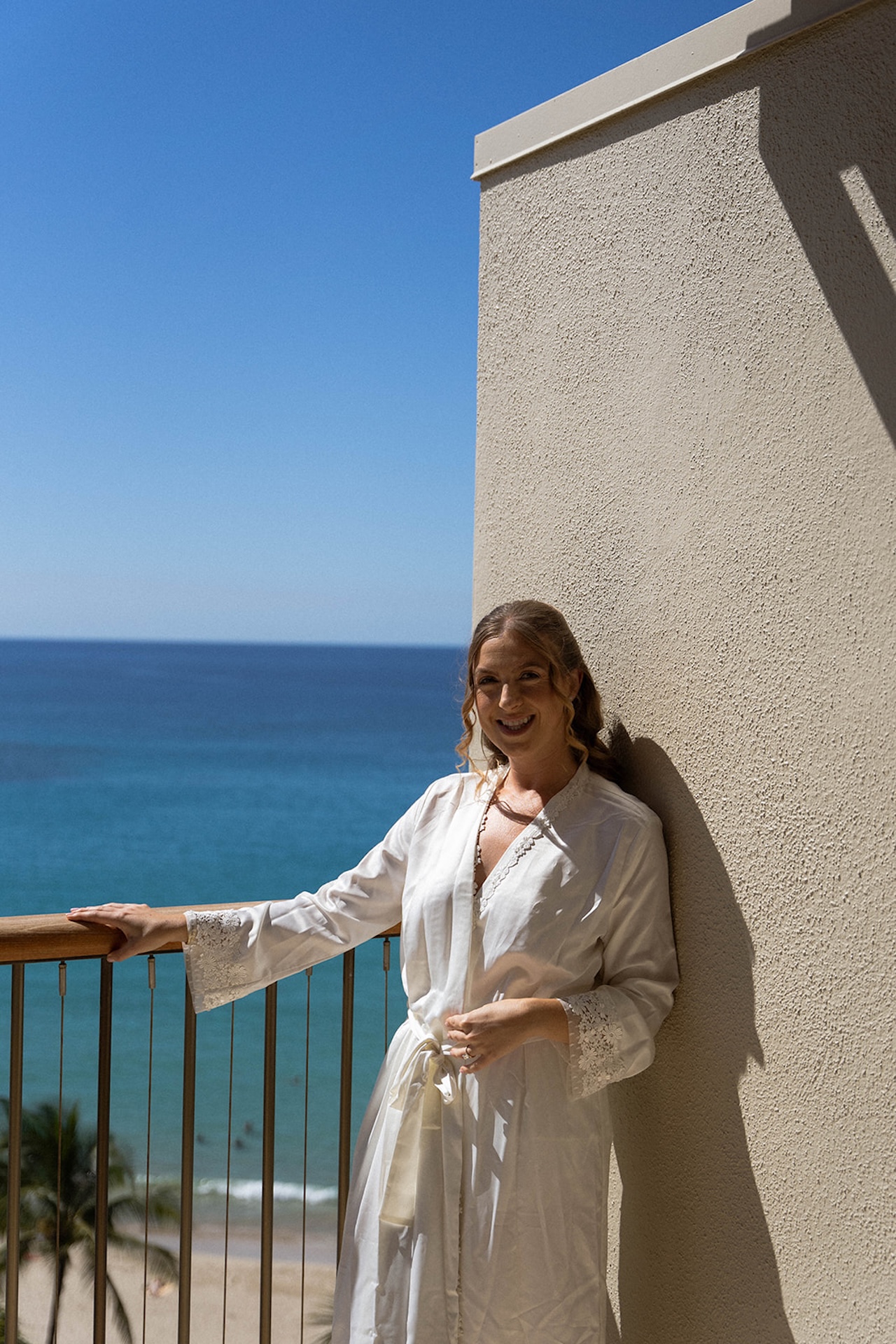 A bride stands on a balcony in a white robe overlooking the ocean, a peaceful morning moment during big island wedding planning.