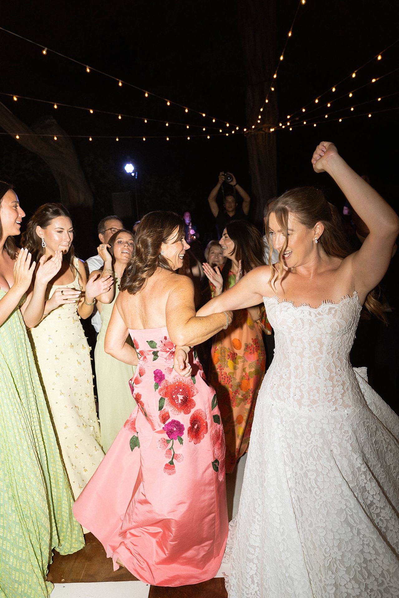 The bride dances joyfully with guests under string lights during a lively outdoor reception.