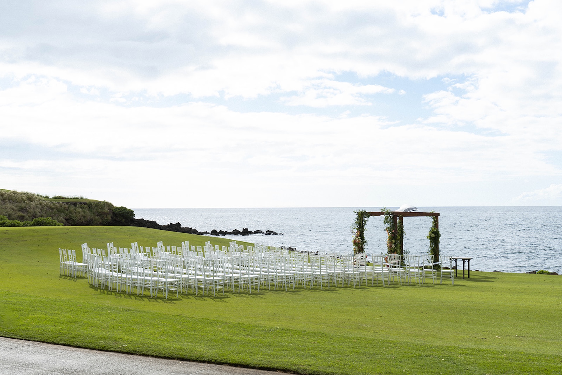 Wide view of an oceanfront wedding ceremony setup with white chairs and a floral arch overlooking the water