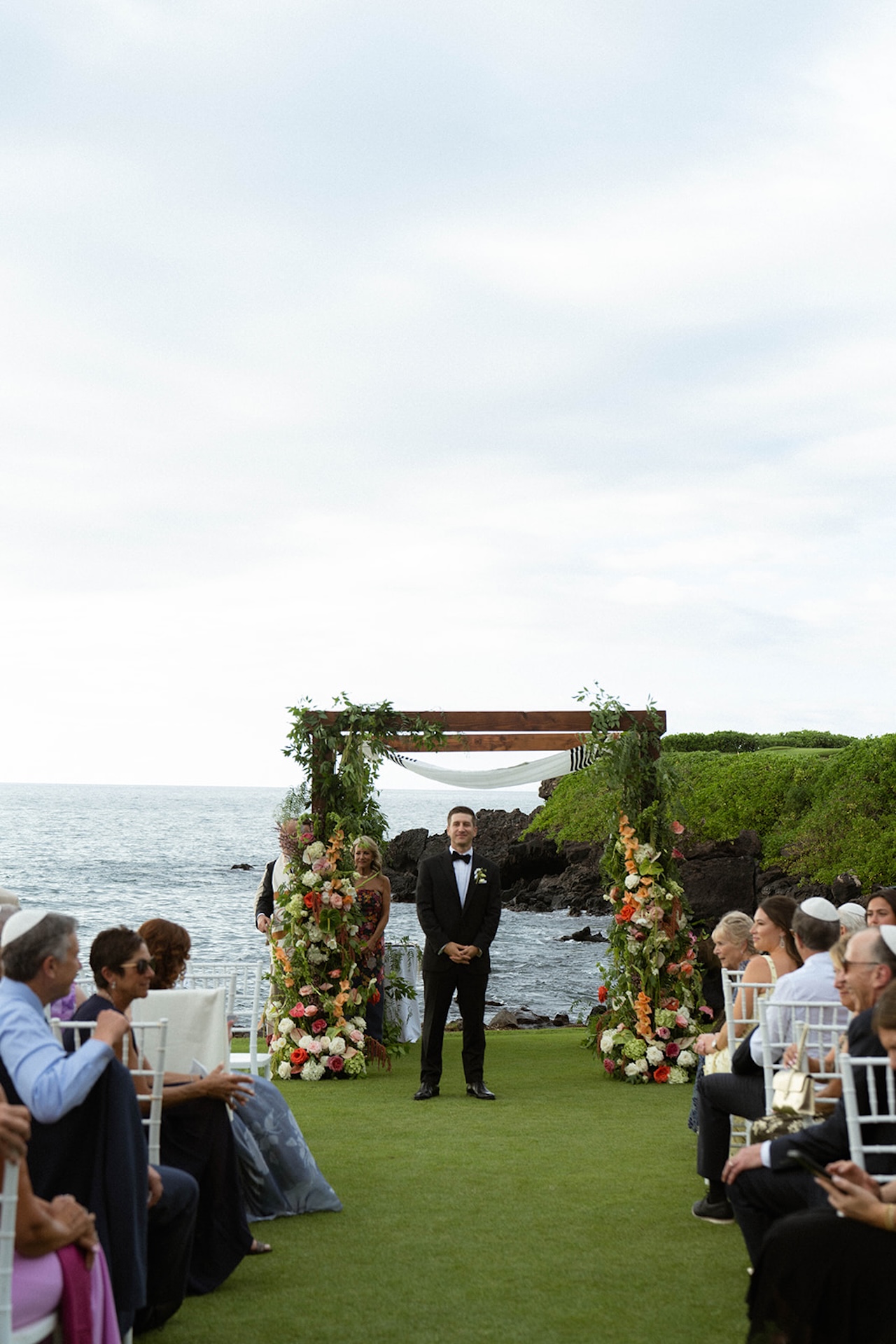 The groom stands beneath a floral arch by the ocean awaiting his partner, a classic scene that reflects thoughtful big island wedding planning.