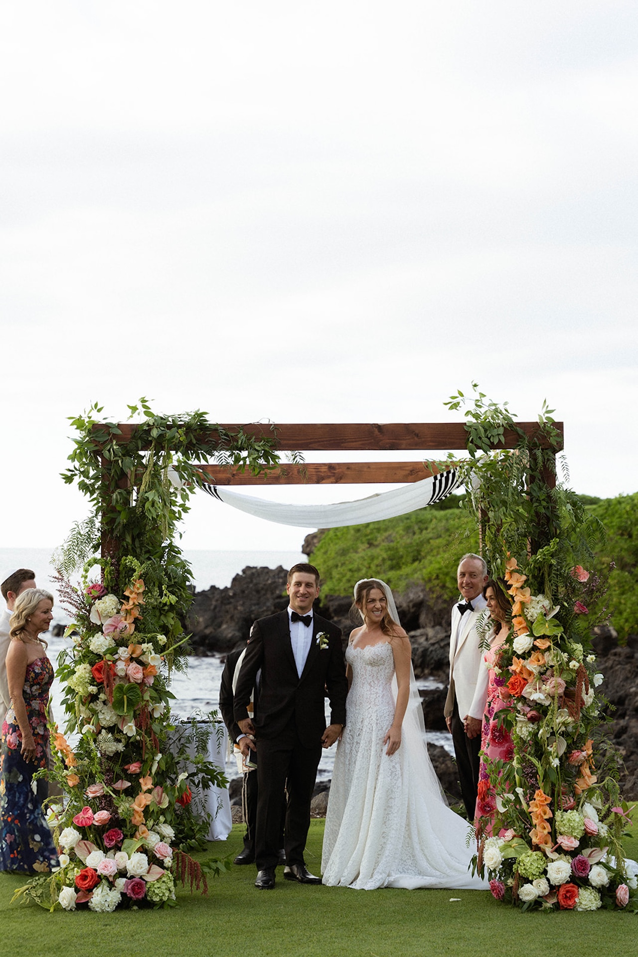 The newly married couple standing beneath a floral ceremony arch by the ocean with family nearby