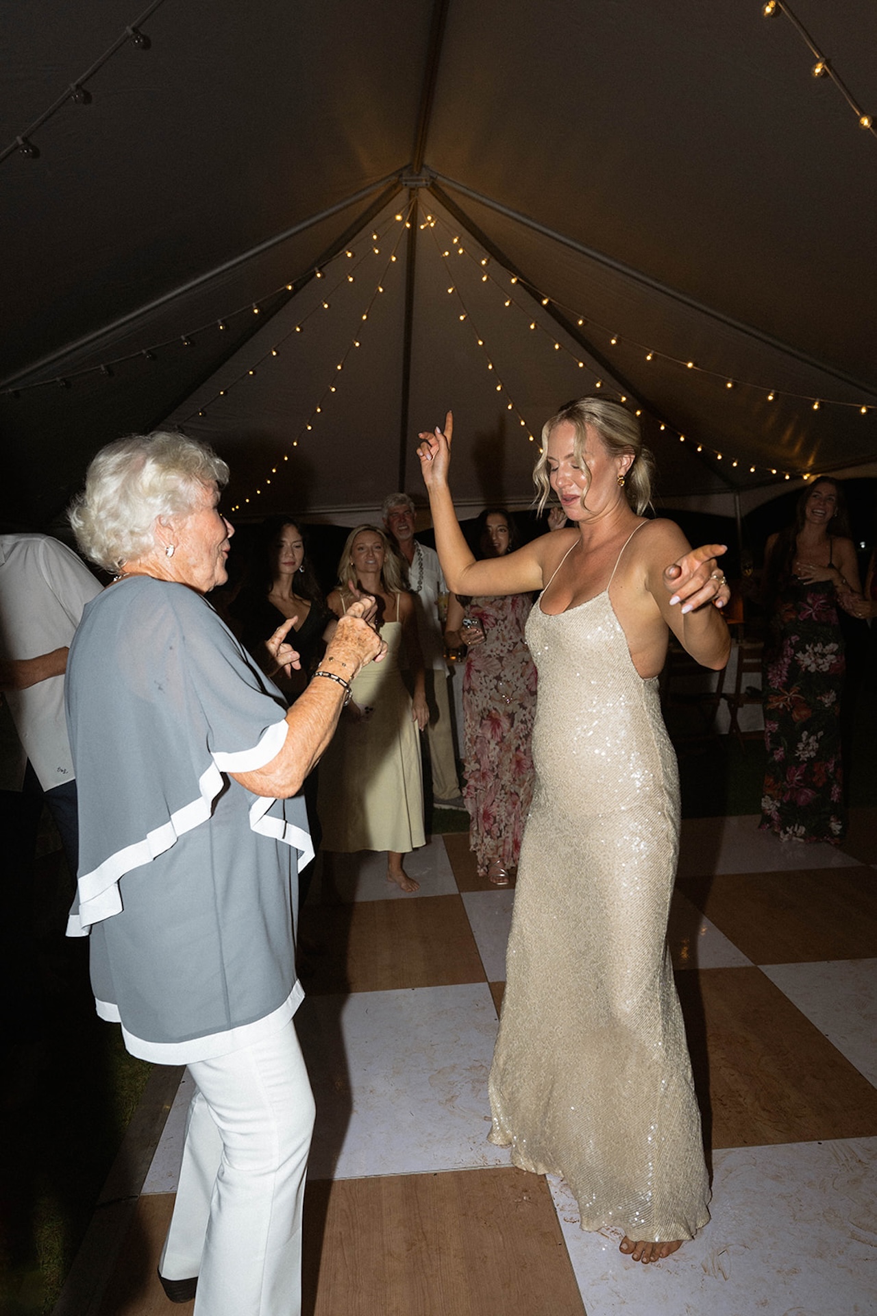 The bride dances joyfully with an older woman under a sailcloth tent as guests cheer during the evening reception.